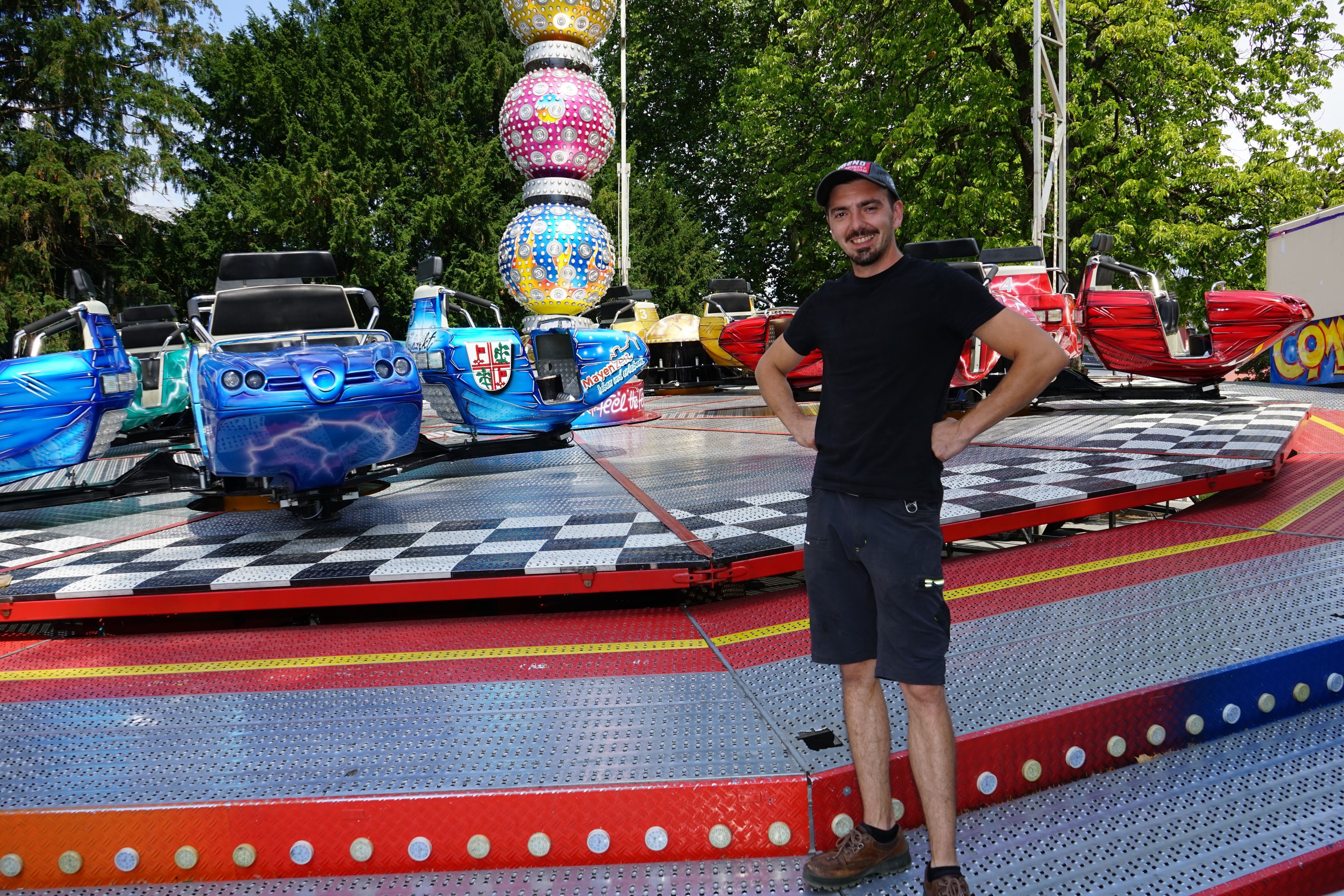 Ein junger Mann steht lächelnd vor einem Kirmes Fahrgeschäft