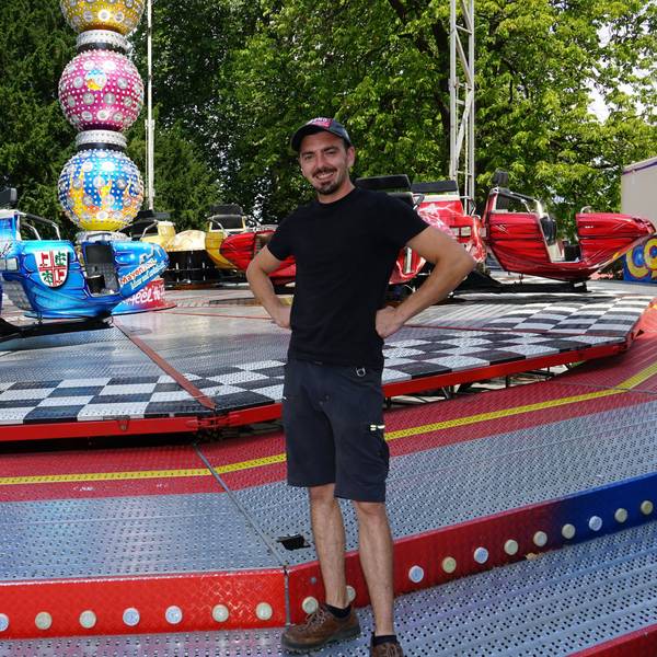 Ein junger Mann steht lächelnd vor einem Kirmes Fahrgeschäft