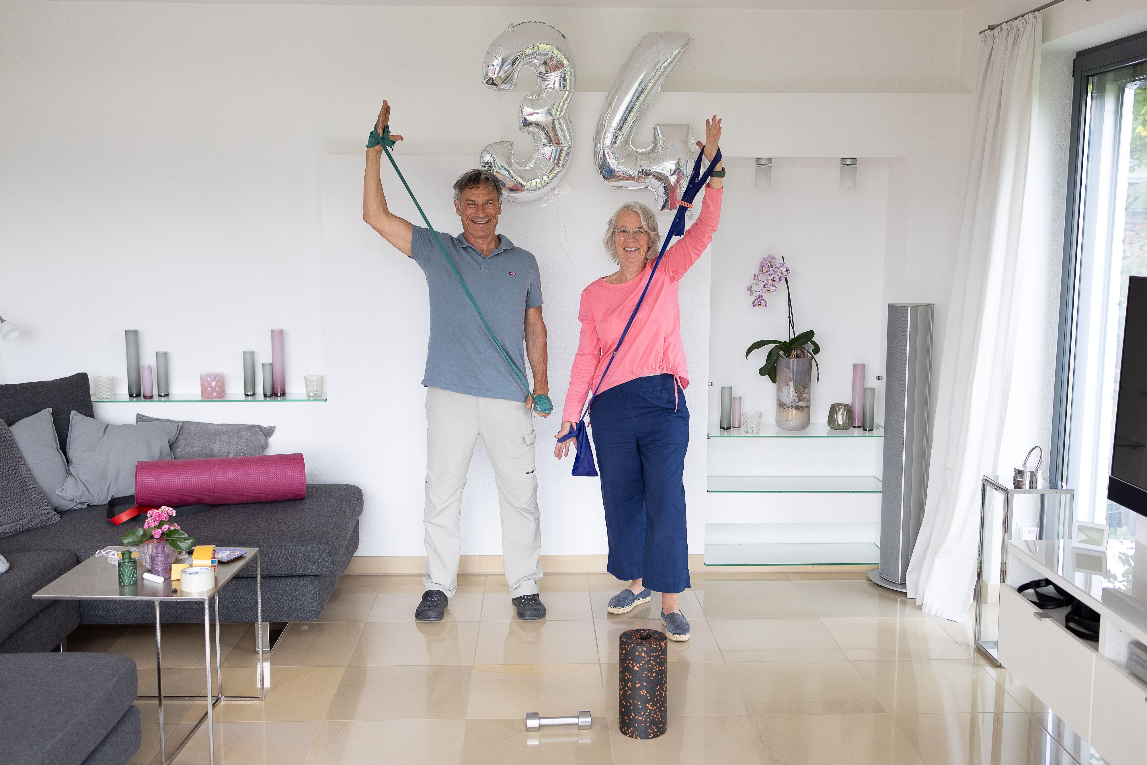 Die Eheleute stehen nebeneinander, sie führen eine Übung mit Therapiebändern aus.
