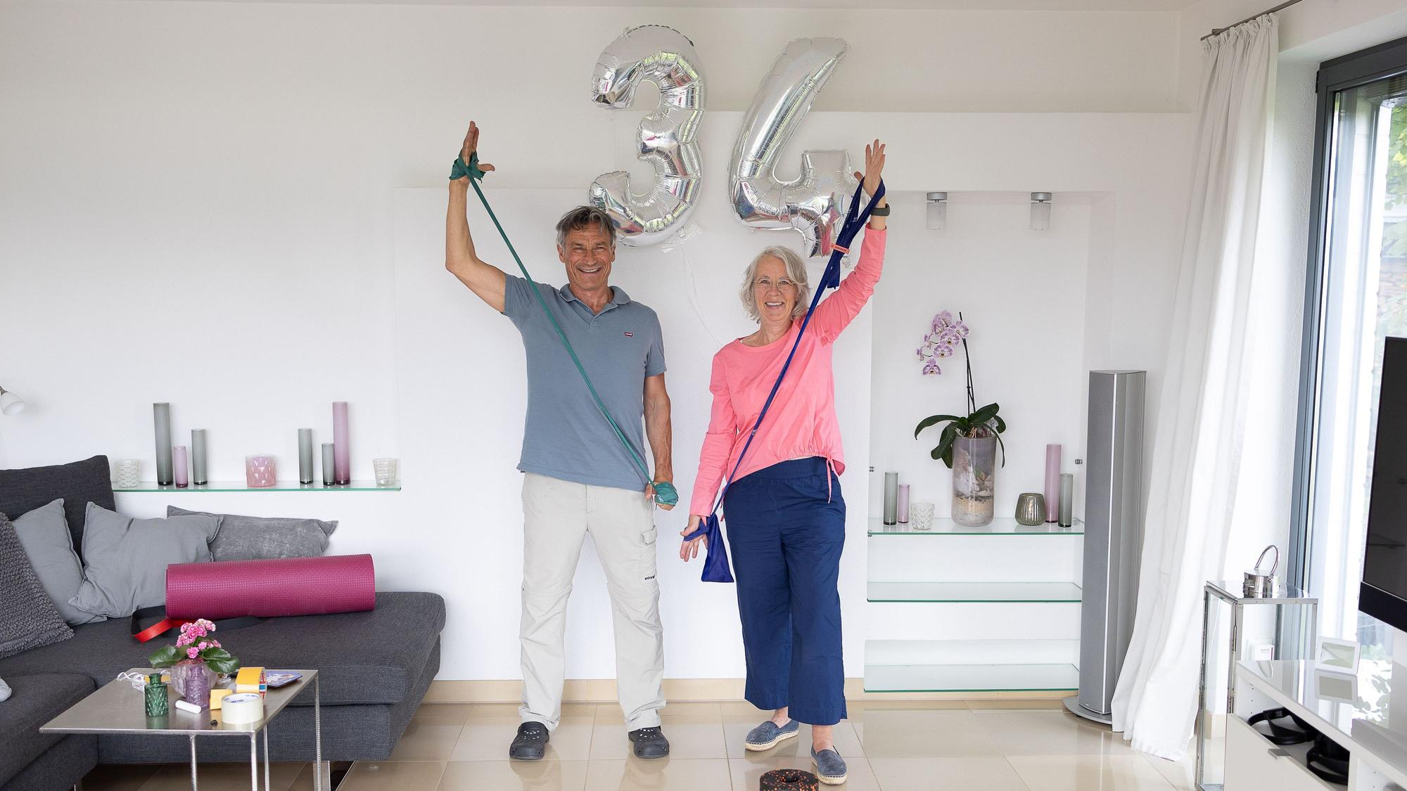 Die Eheleute stehen nebeneinander, sie führen eine Übung mit Therapiebändern aus.