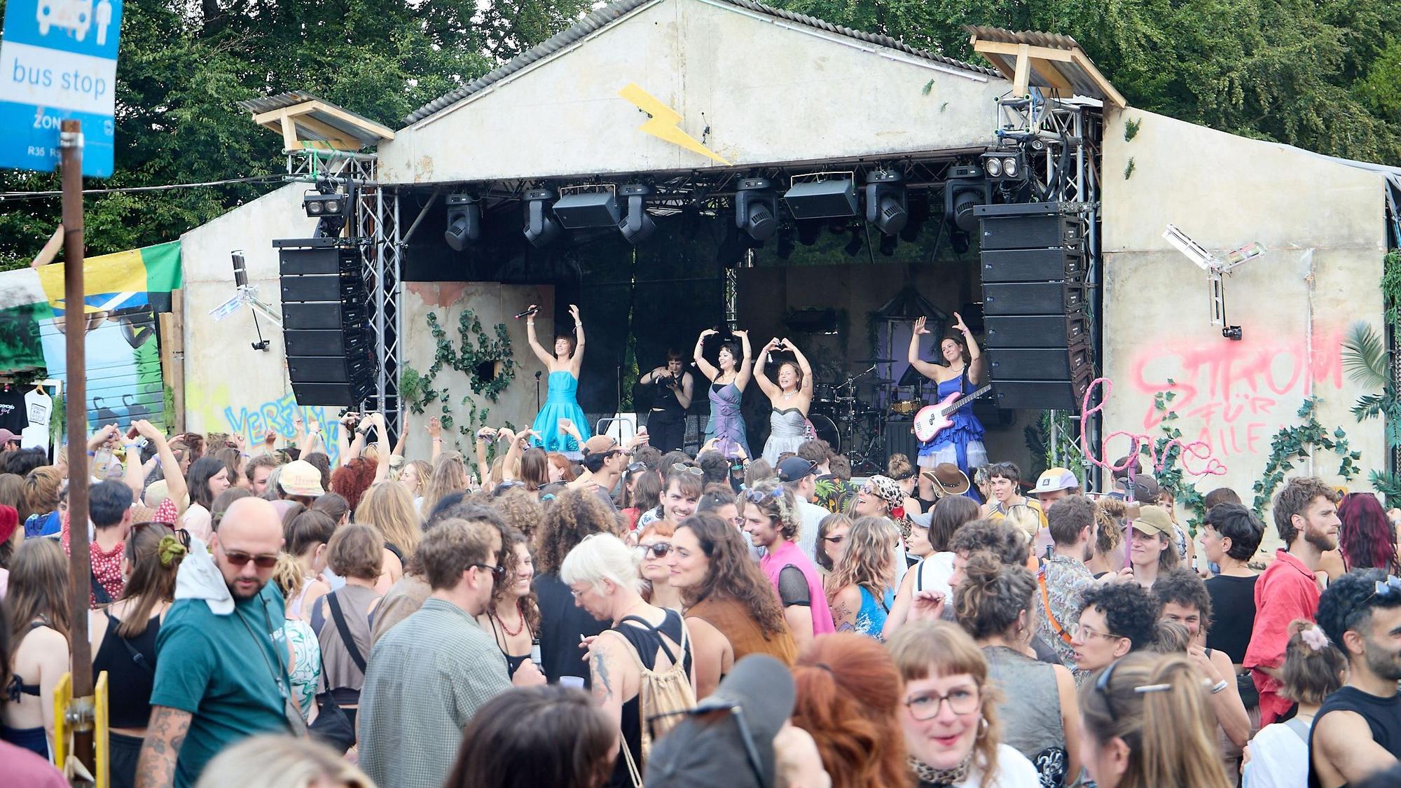 Bandmitglieder heben auf der Bühne des Festivals die Arme, davor stehen die Zuschauer.