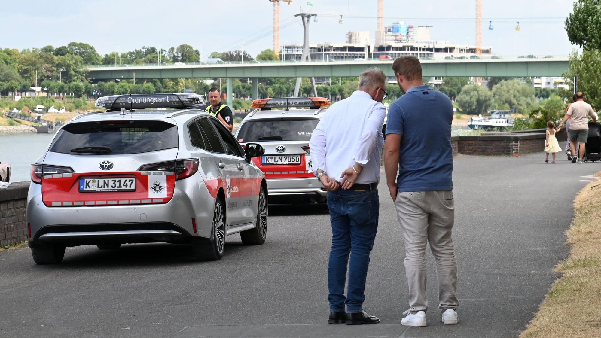 Zwei Männer stehen mit dem Rücken zur Kamera. Daneben ein Auto vom Ordnungsamt.