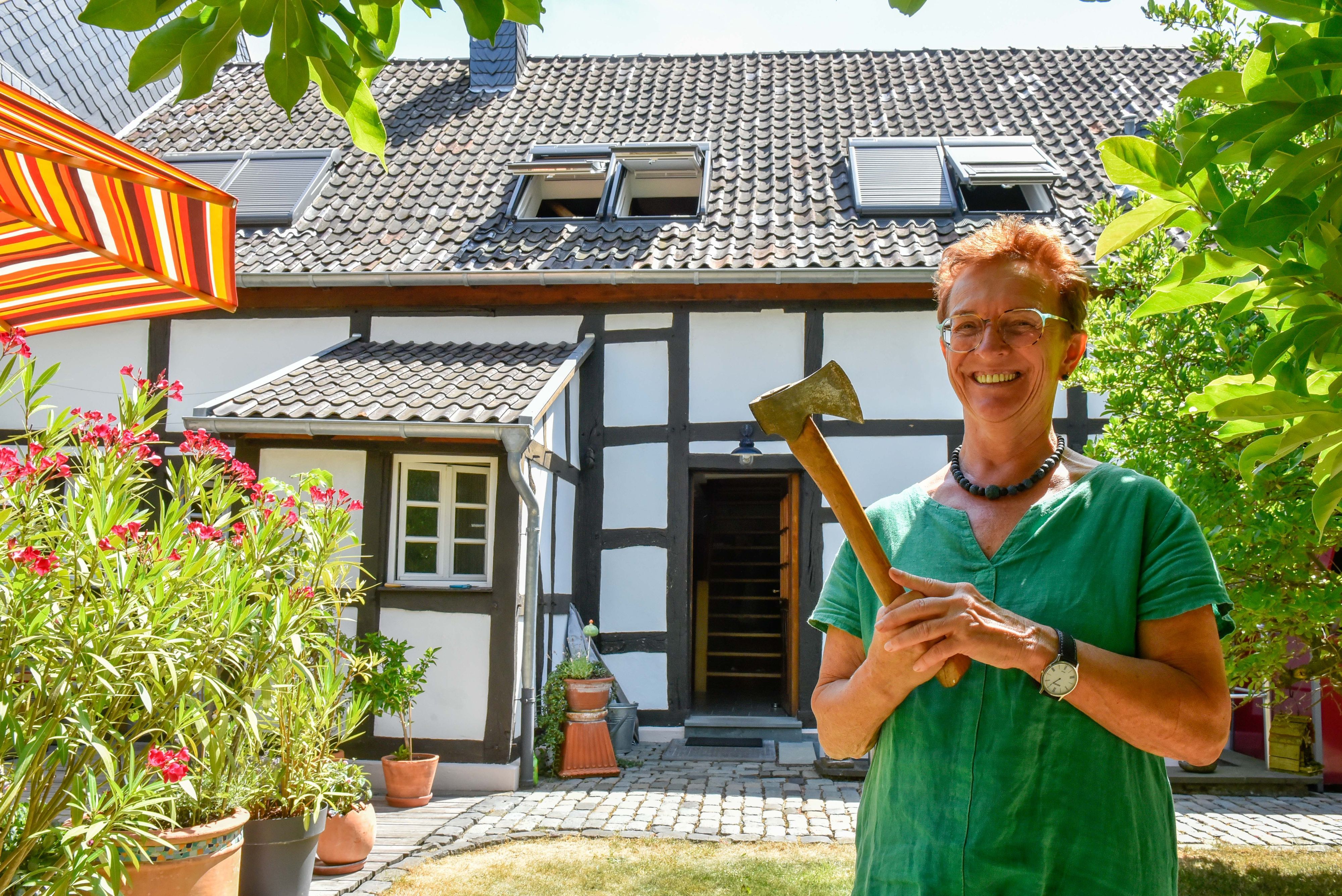 Eine lächelnde Frau im grünen Shirt steht mit einem Beil in der Hand vor einem restaurierten Bauernhof.
