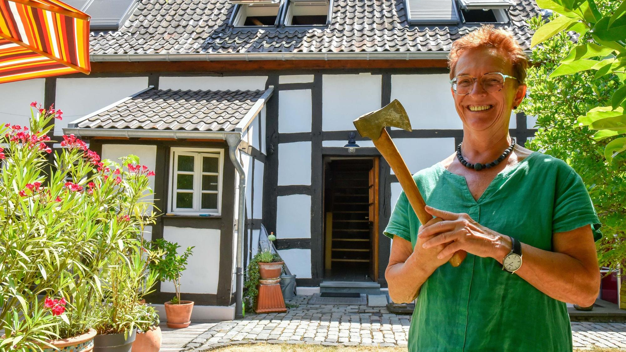 Eine lächelnde Frau im grünen Shirt steht mit einem Beil in der Hand vor einem restaurierten Bauernhof.