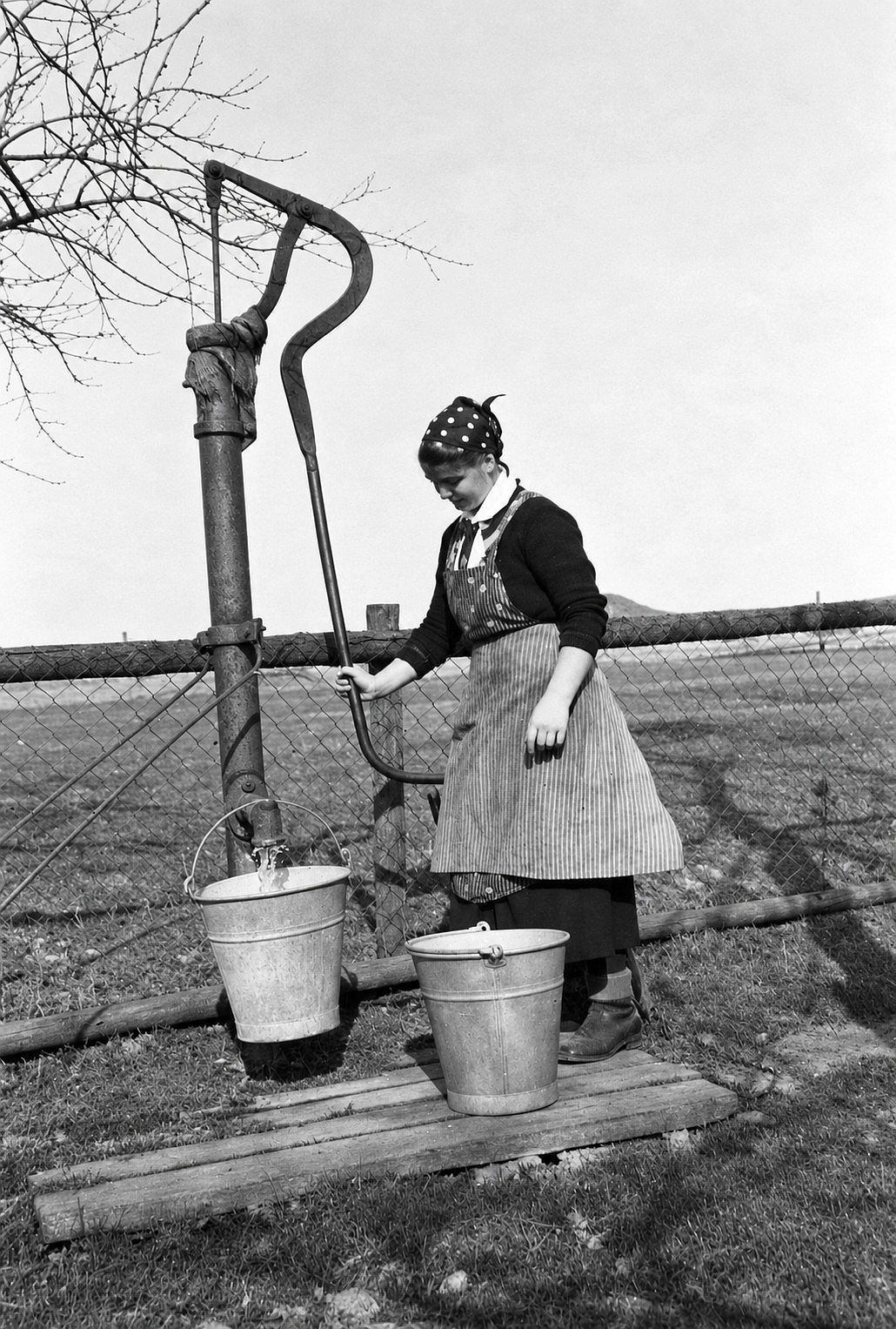 Eine Frau schöpft in den 1930er Jahren in Morsbach mit einer Schwengelpumpe Wasser aus einem Brunnen in zwei Zinkeimer.