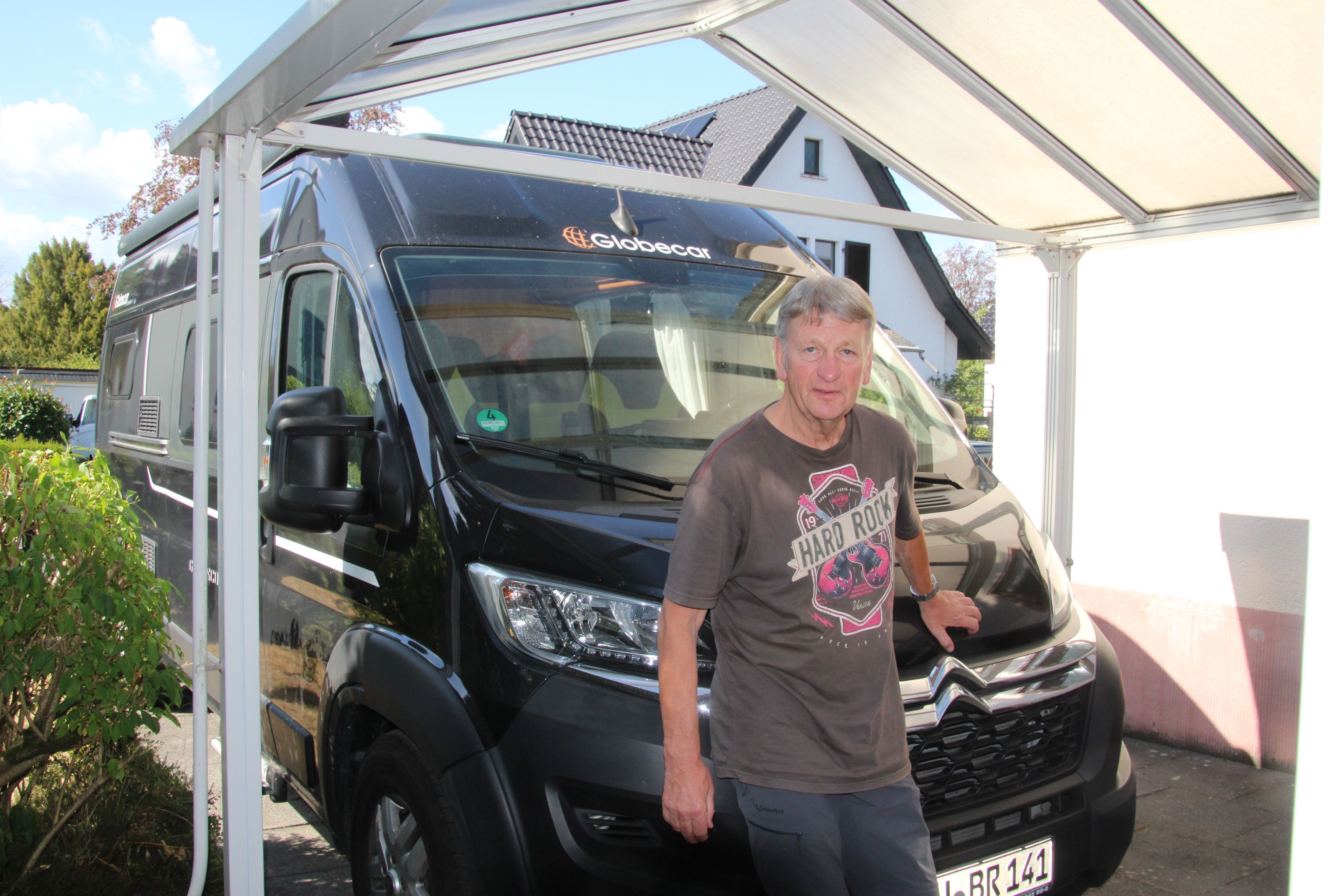 Ein Mann in einem bedruckten T-Shirt steht unter einem Carport vor einem Wohnmobil.