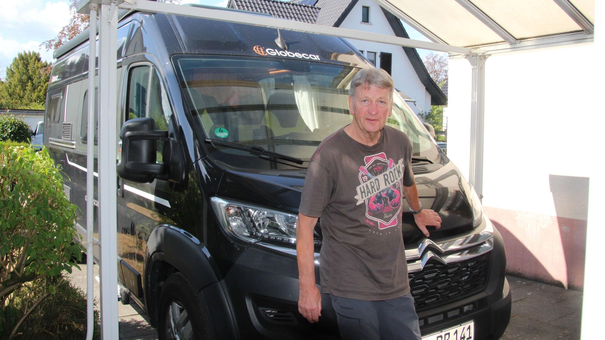 Ein Mann in einem bedruckten T-Shirt steht unter einem Carport vor einem Wohnmobil.