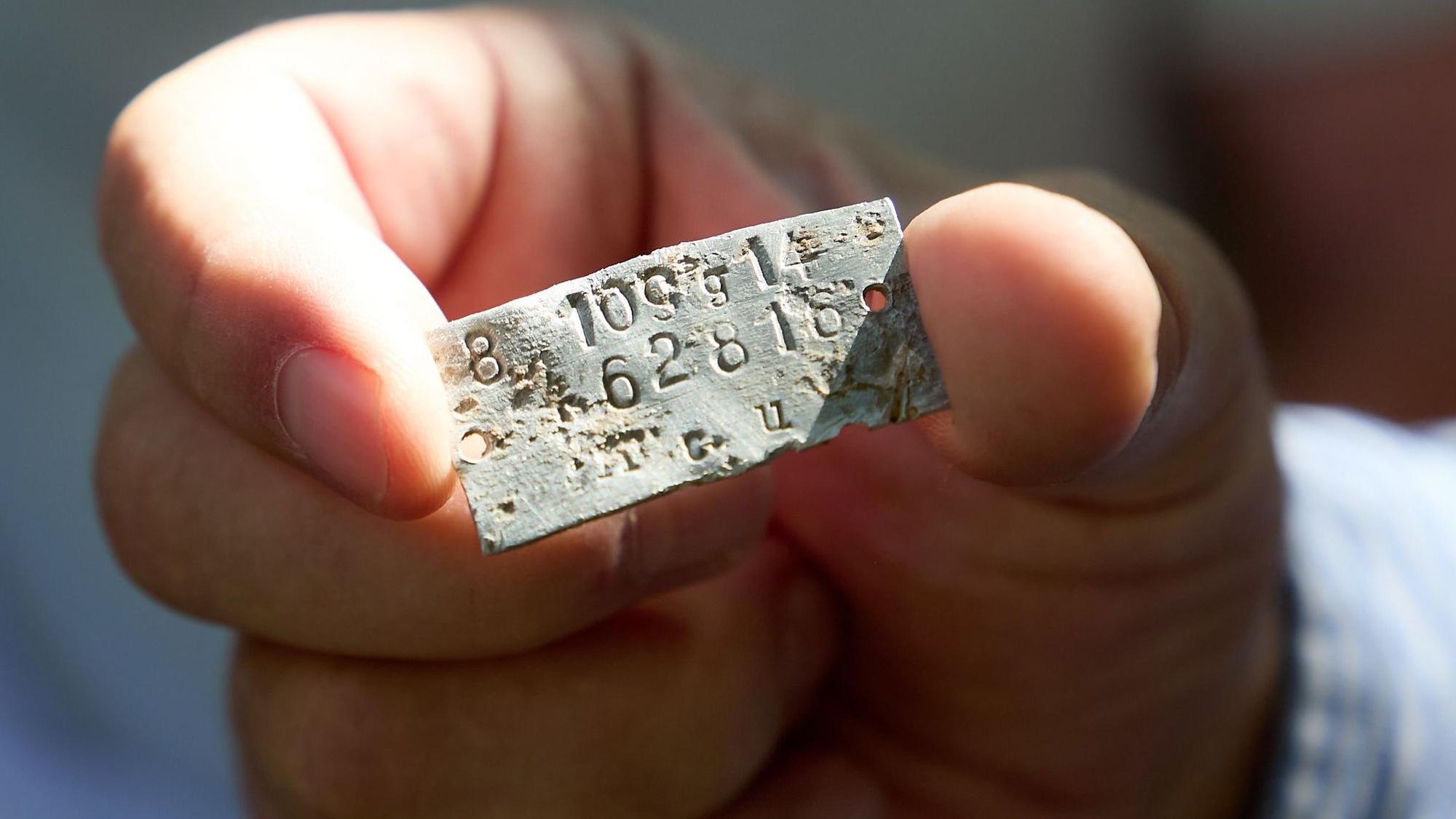 Ein Mann hällt ein Metallplättchen, auf dem Zahlen stehen, zwischen Daumen und Zeigefinger.