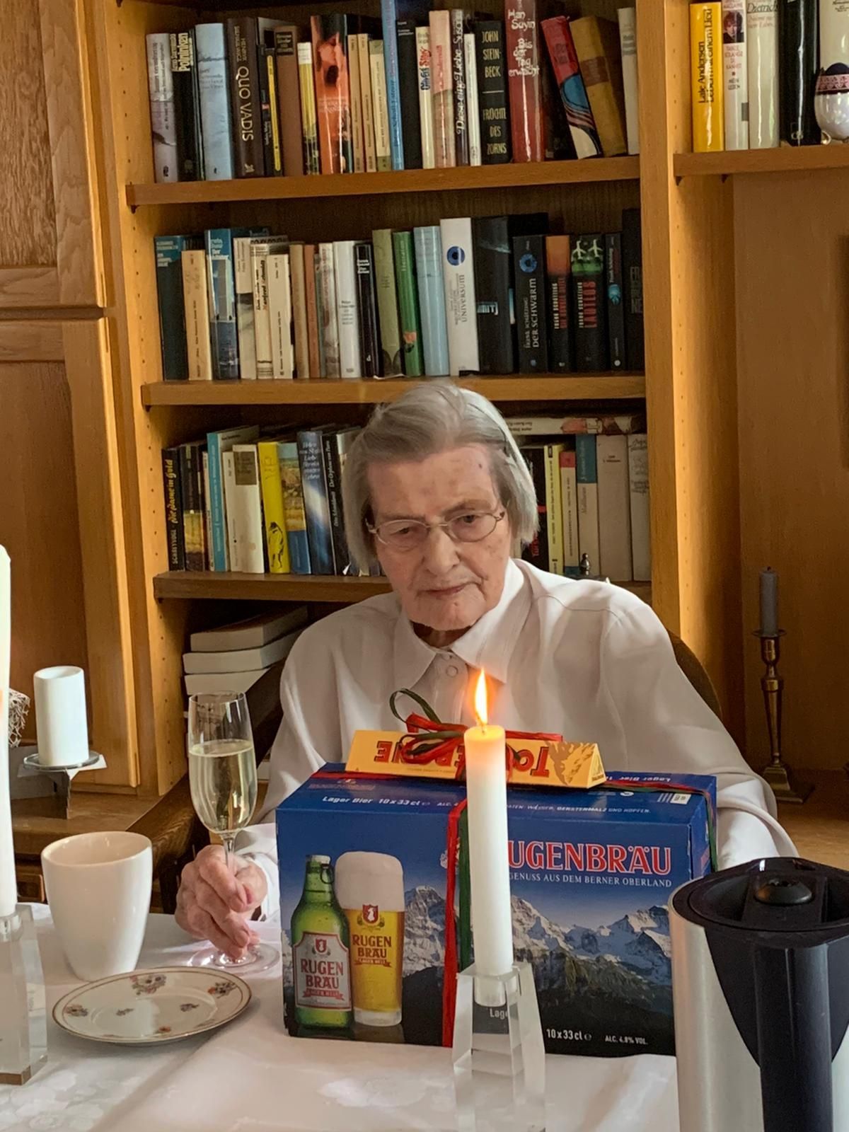 Das Foto zeigt eine ältere Frau mit einem Sektglas in der Hand, vor ihr eine Kerze und eine Packung Bier als Geschenk verpackt.