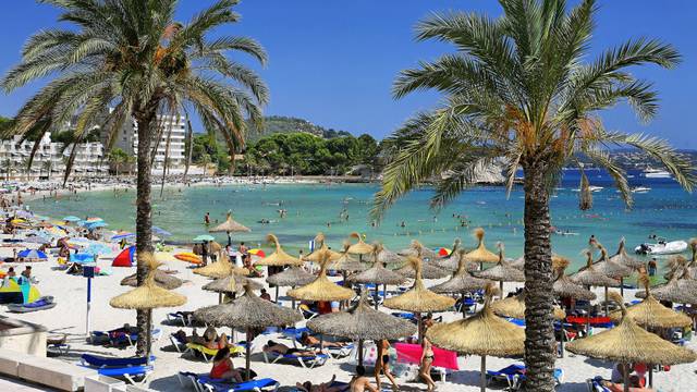 Urlauber an einer Badebucht mit Sonnenschirmen und Palmen.