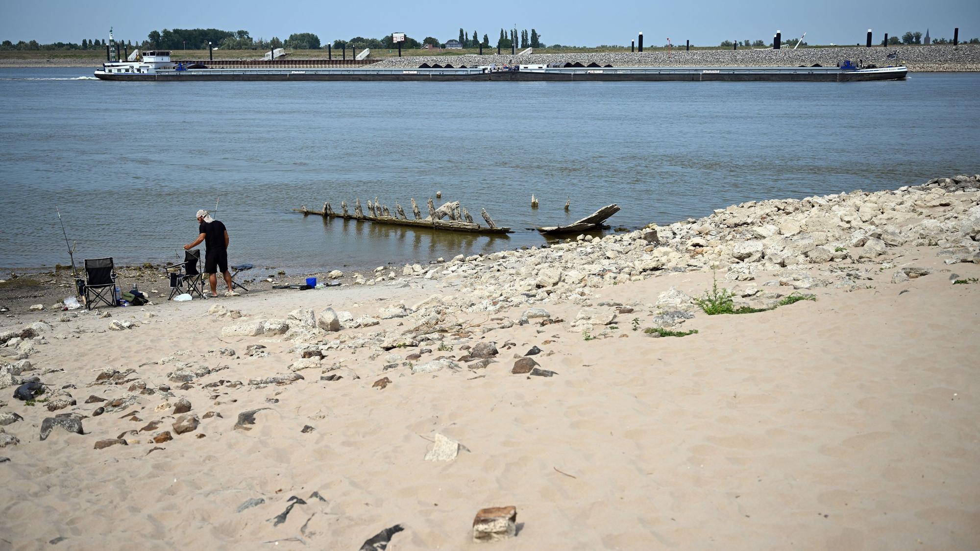 Ein Frachtschiff fährt bei Niedrigwasser auf dem Rhein an dem Wrack eines Bootes vorbei, das normalerweise unter Wasser liegt.