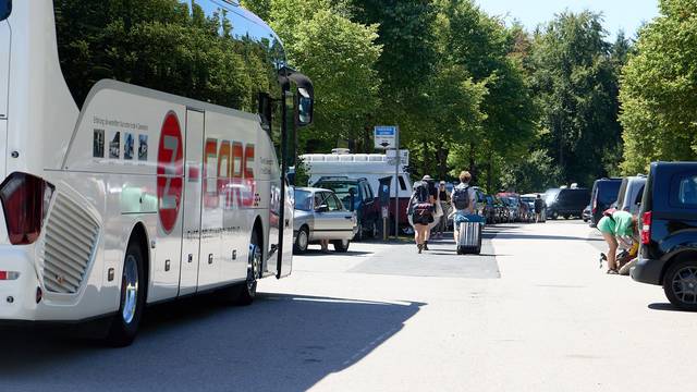 Menschen laufen bepackt über einen Parkplatz, auf dem Busse und Autos parken.