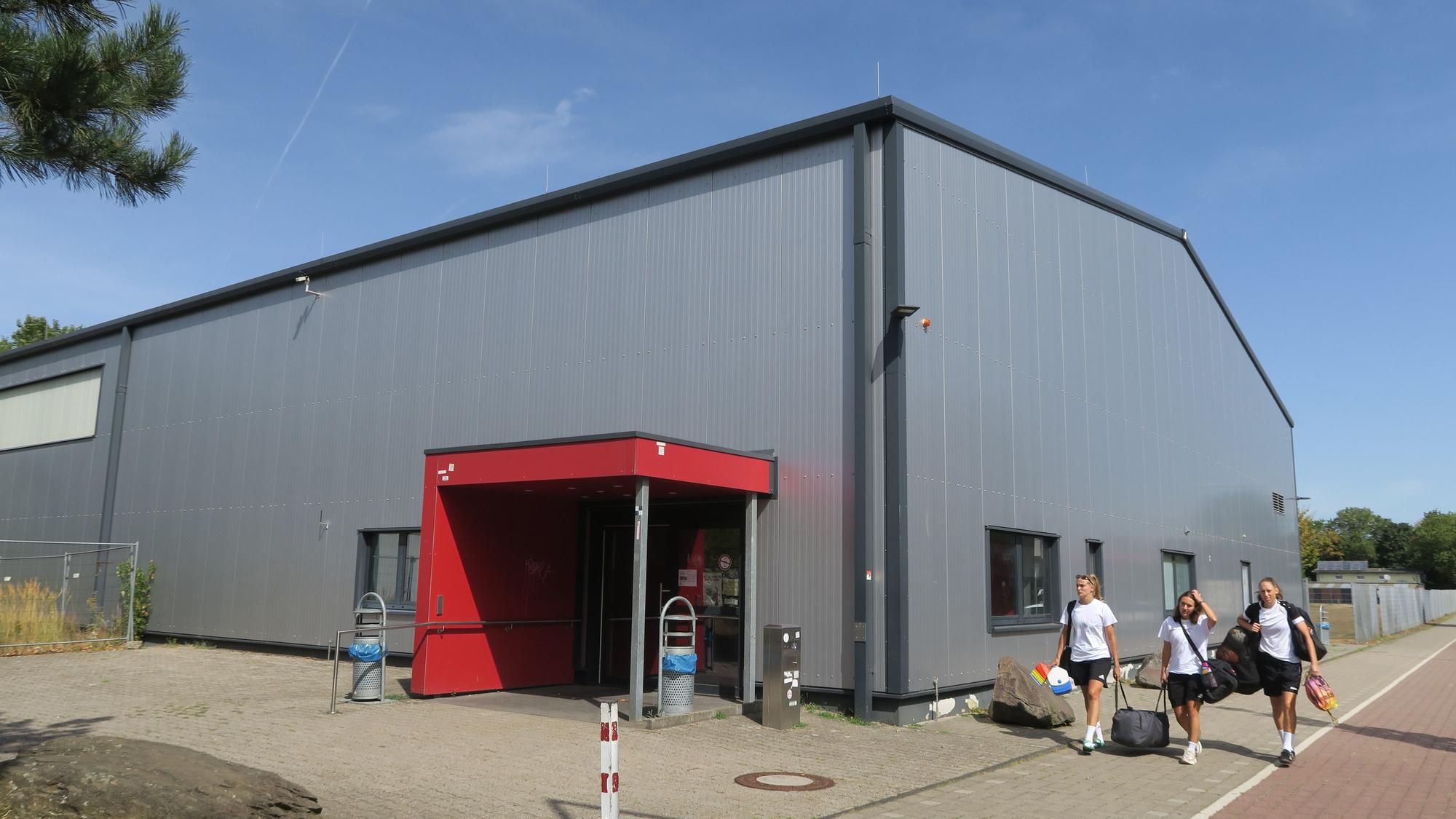 Eine silberfarbene Sporthalle ist zu sehen. Drei sportlich gekleidete Menschen laufen an ihr vorbei.