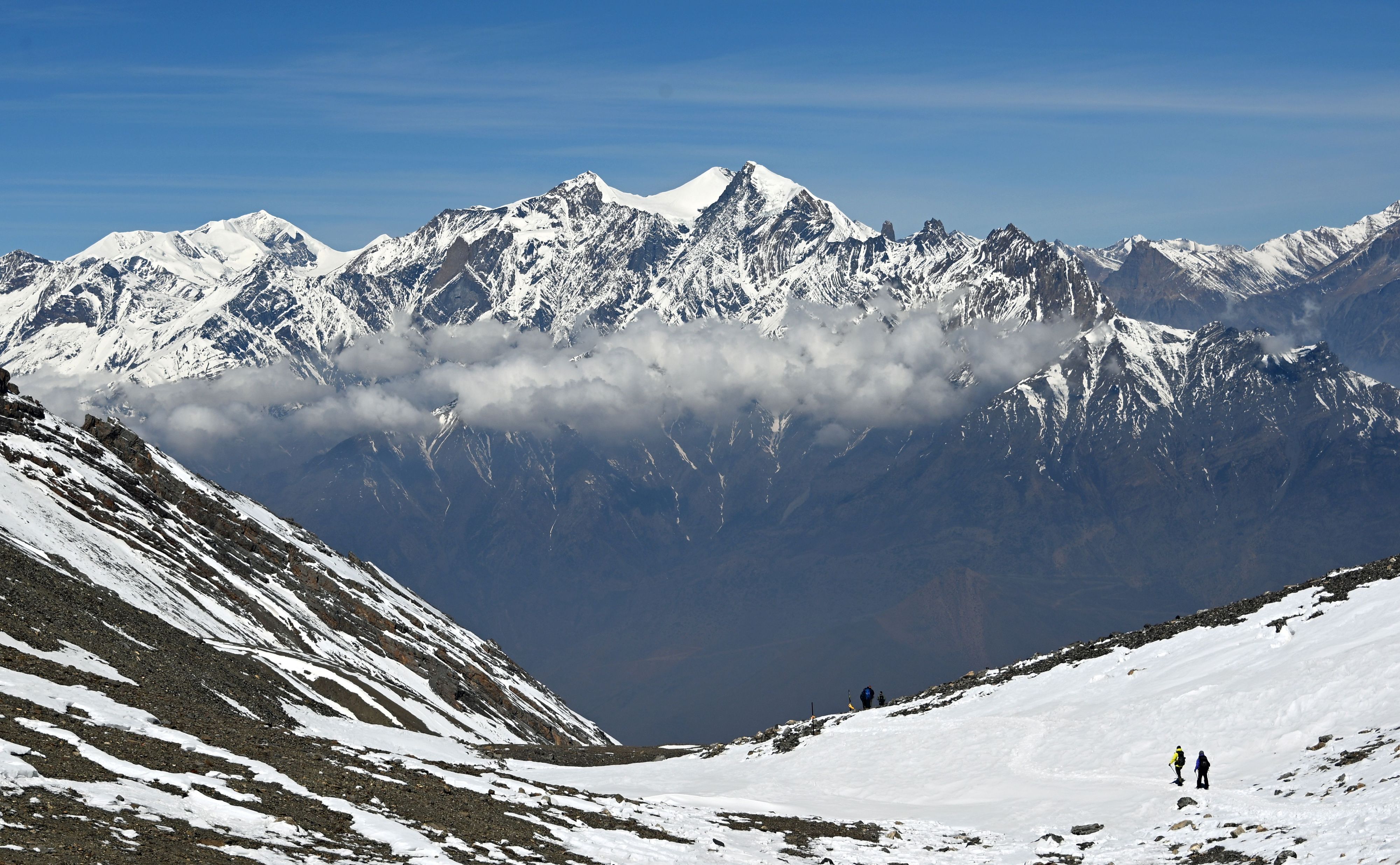Wanderer gehen am Thorong La Pass in der Annapurna-Region auf einem schneebedeckten Weg, im Hintergrund ragen schneebedeckte Berggipfel des Himalaya über einer Wolkenschicht auf.