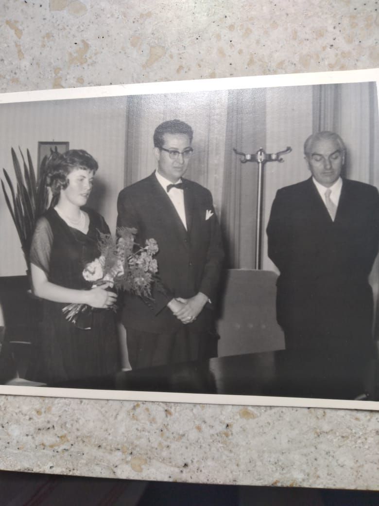 Das Bild zeigt eine Frau mit Blumenstrauß und zwei Männer in Anzügen in Schwarz-Weiß.