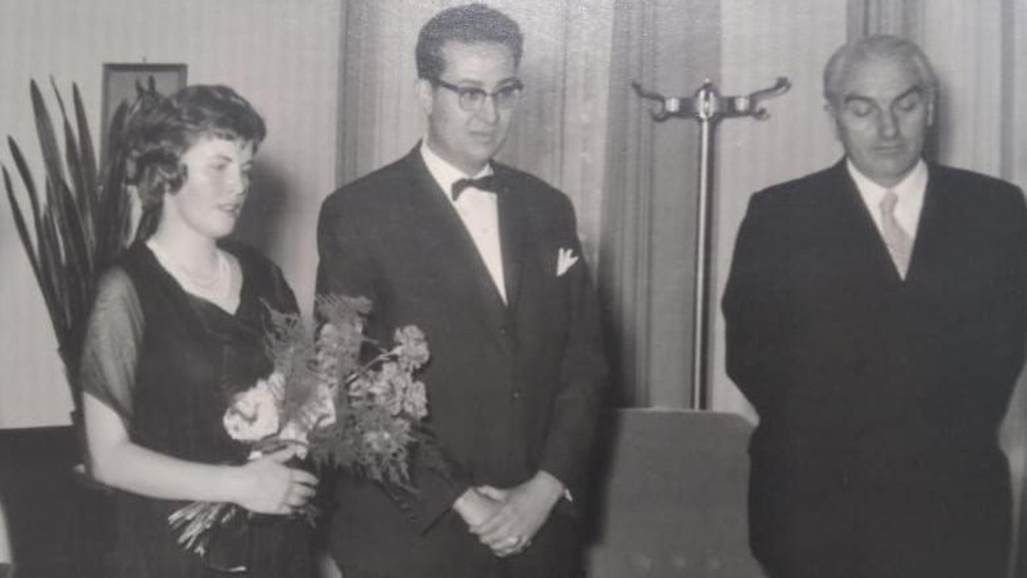 Das Bild zeigt eine Frau mit Blumenstrauß und zwei Männer in Anzügen in Schwarz-Weiß.