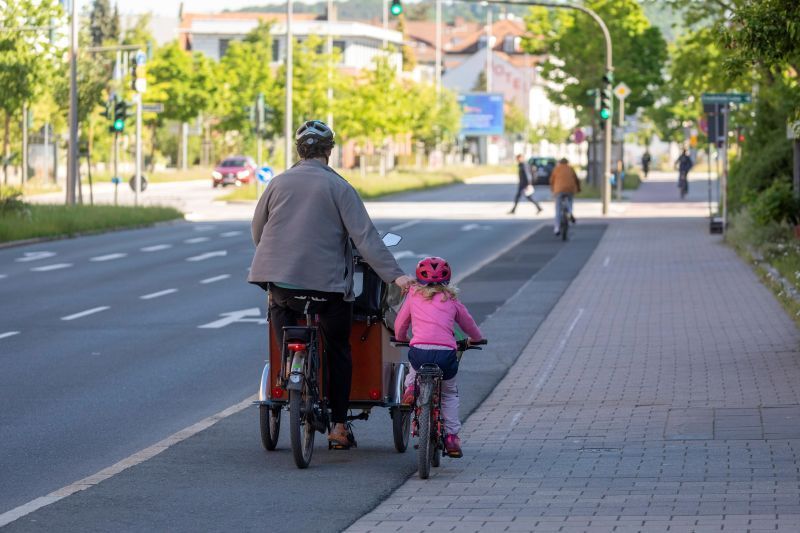 Ein Mann fährt mit einem Lastenrad, daneben fährt ein Kind auf einem Kinderrad.