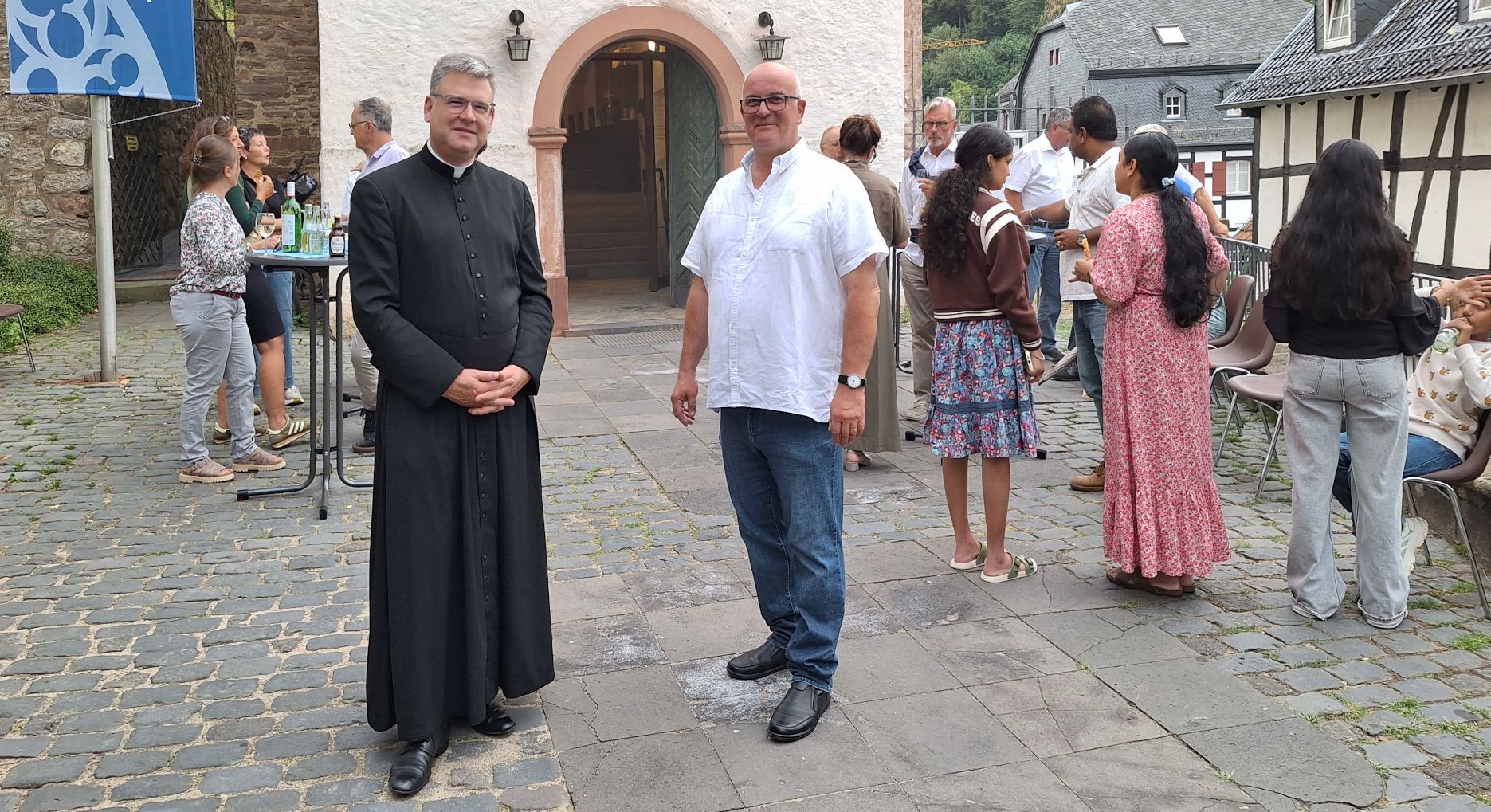 Ein Pfarrer und ein anderer Mann stehen vor dem Eingang zur Kirche. Im Hintergrund sind weitere Männer und Frauen zu sehen.