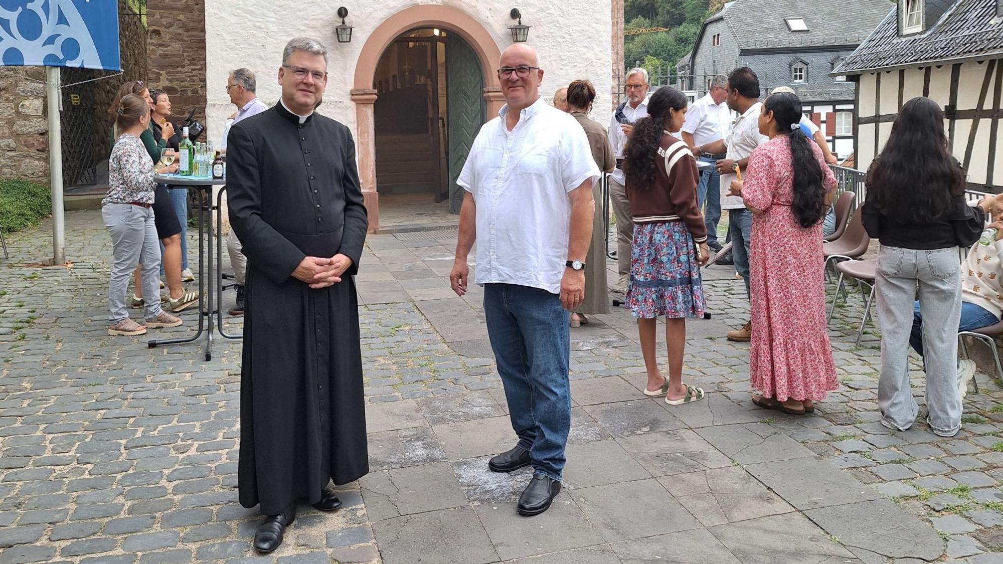 Ein Pfarrer und ein anderer Mann stehen vor dem Eingang zur Kirche. Im Hintergrund sind weitere Männer und Frauen zu sehen.
