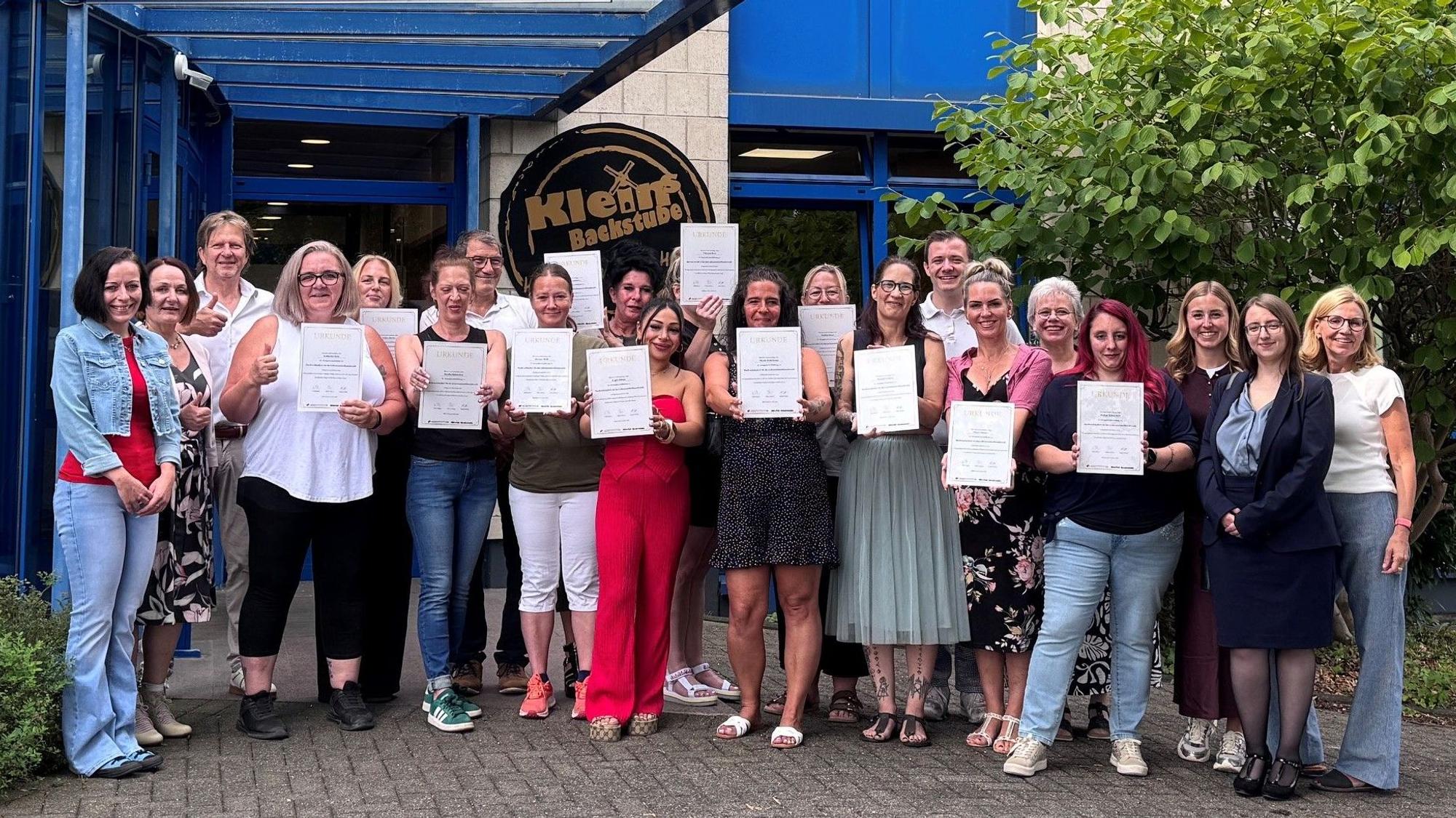 Das Foto zeigt die Absolventinnen mit Urkunden sowie Gratulanten vor der Firmenzentale von „Klein's Backstube“ in Hürth.