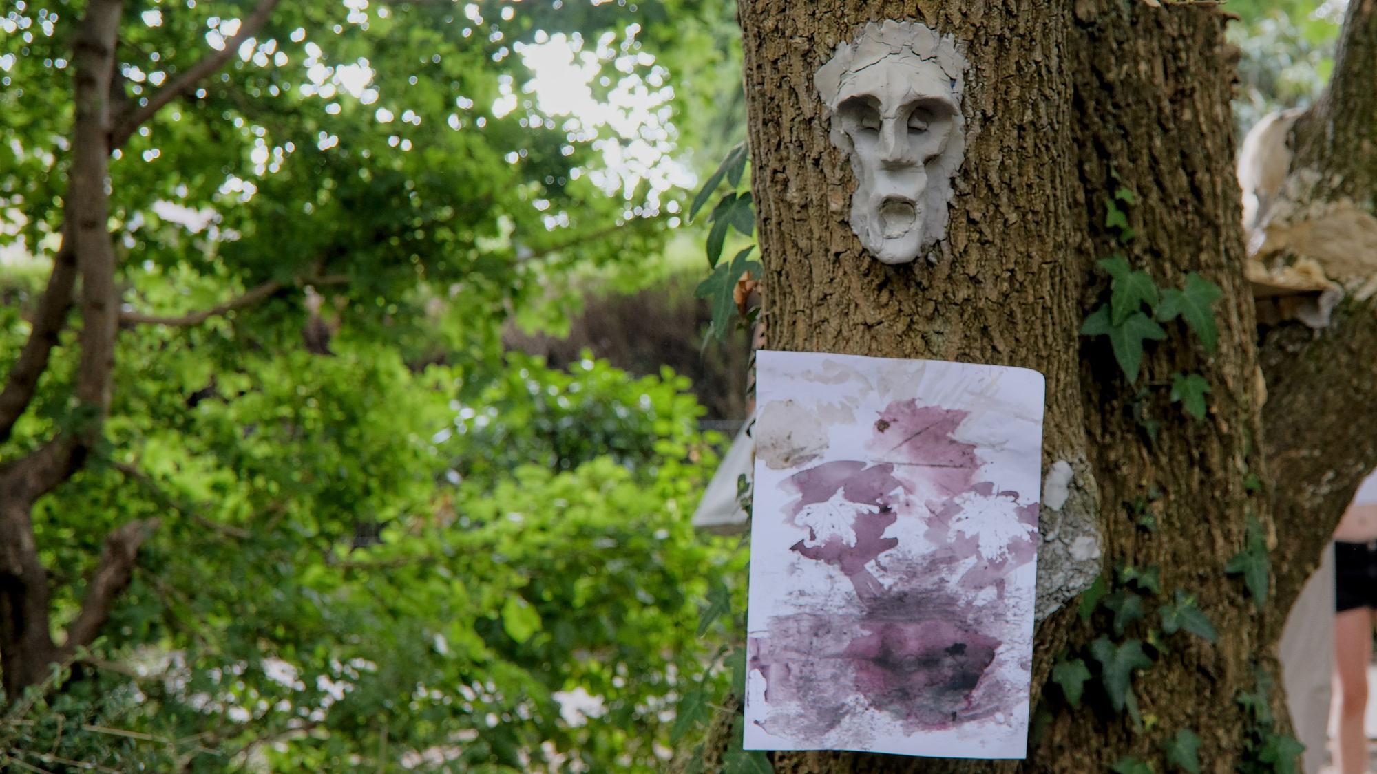 An einem Baumstamm hängt ein Poster mit Farbkleksen udn darüber eine menschliche Maske aus Ton.