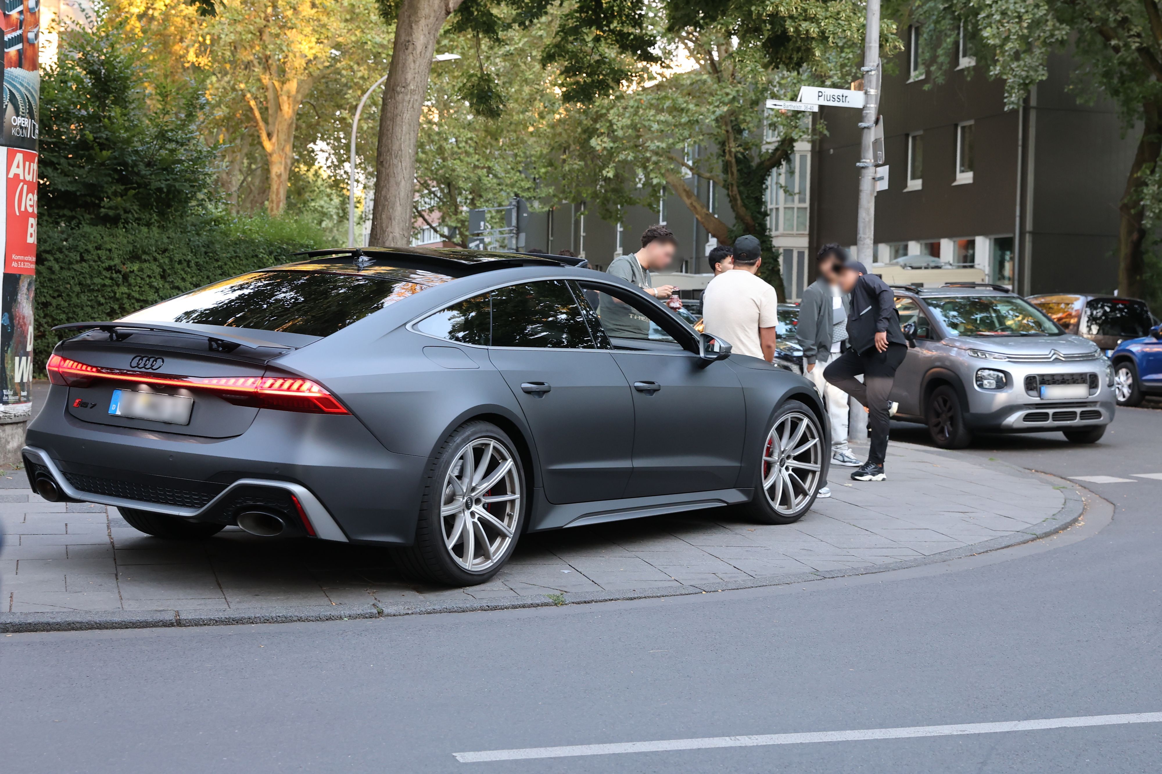 Ein Audi parkt auf dem Bürgersteig an einer Straßenecke. Fünf junge Leute stehen daneben.