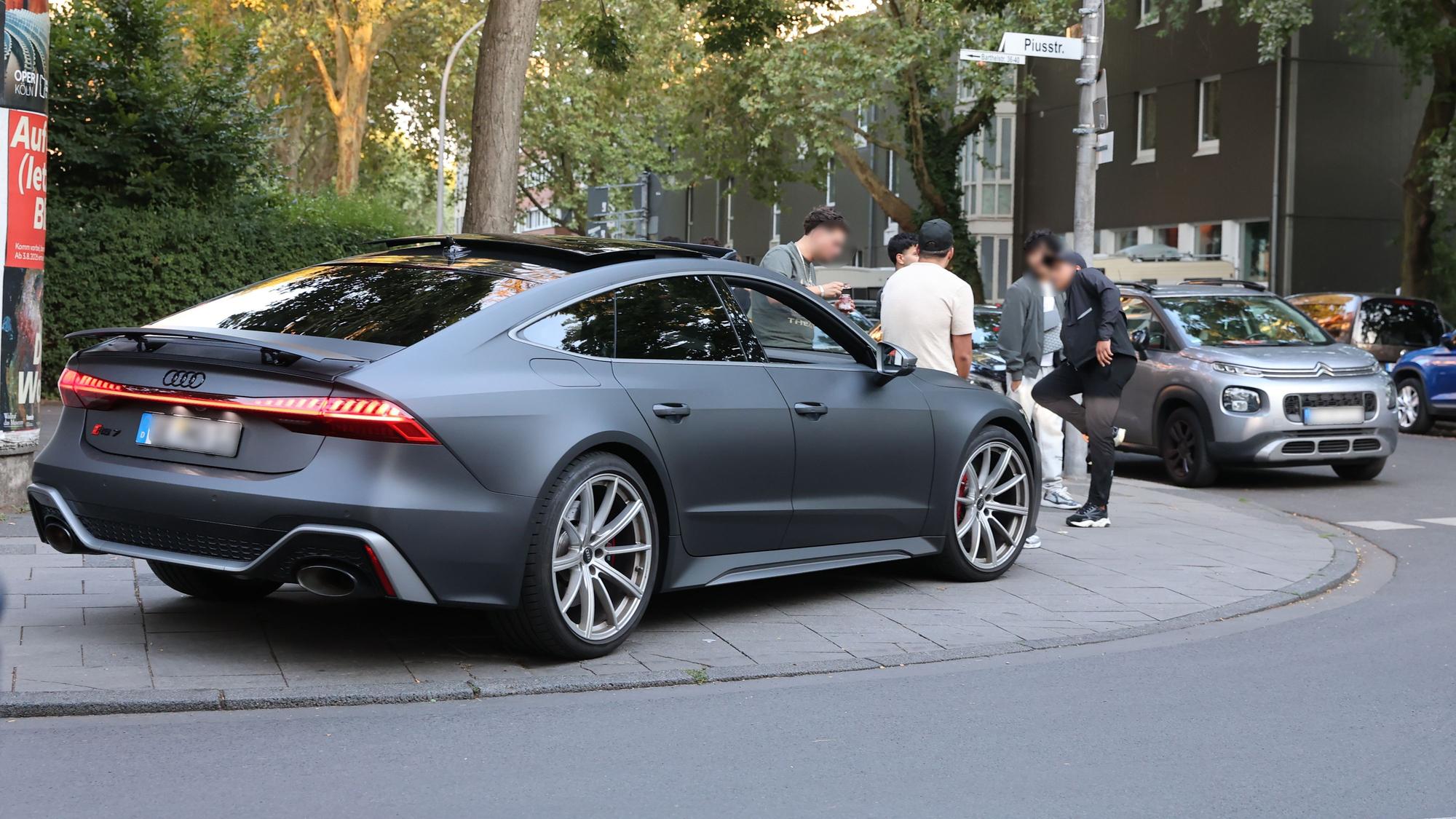 Ein Audi parkt auf dem Bürgersteig an einer Straßenecke. Fünf junge Leute stehen daneben.