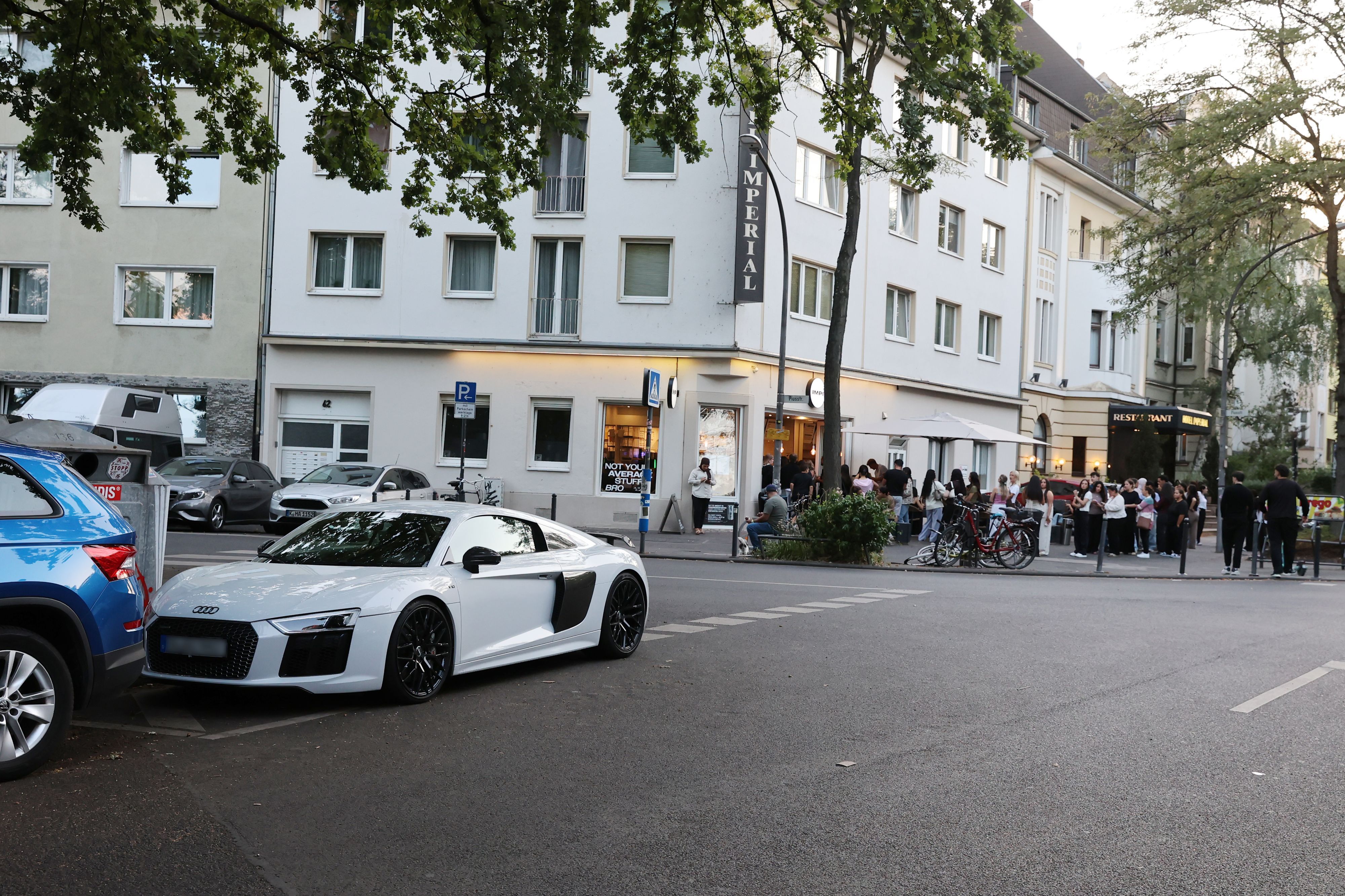 An einer Straßenkreuzung stehen Menschen Schlange vor einem Café. An der Ecke parkt ein PS-starkes Auto verkehrswidrig.
