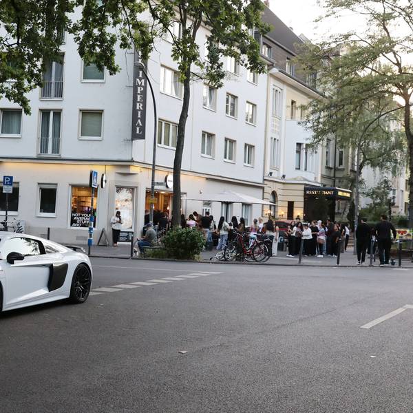 An einer Straßenkreuzung stehen Menschen Schlange vor einem Café. An der Ecke parkt ein PS-starkes Auto verkehrswidrig.