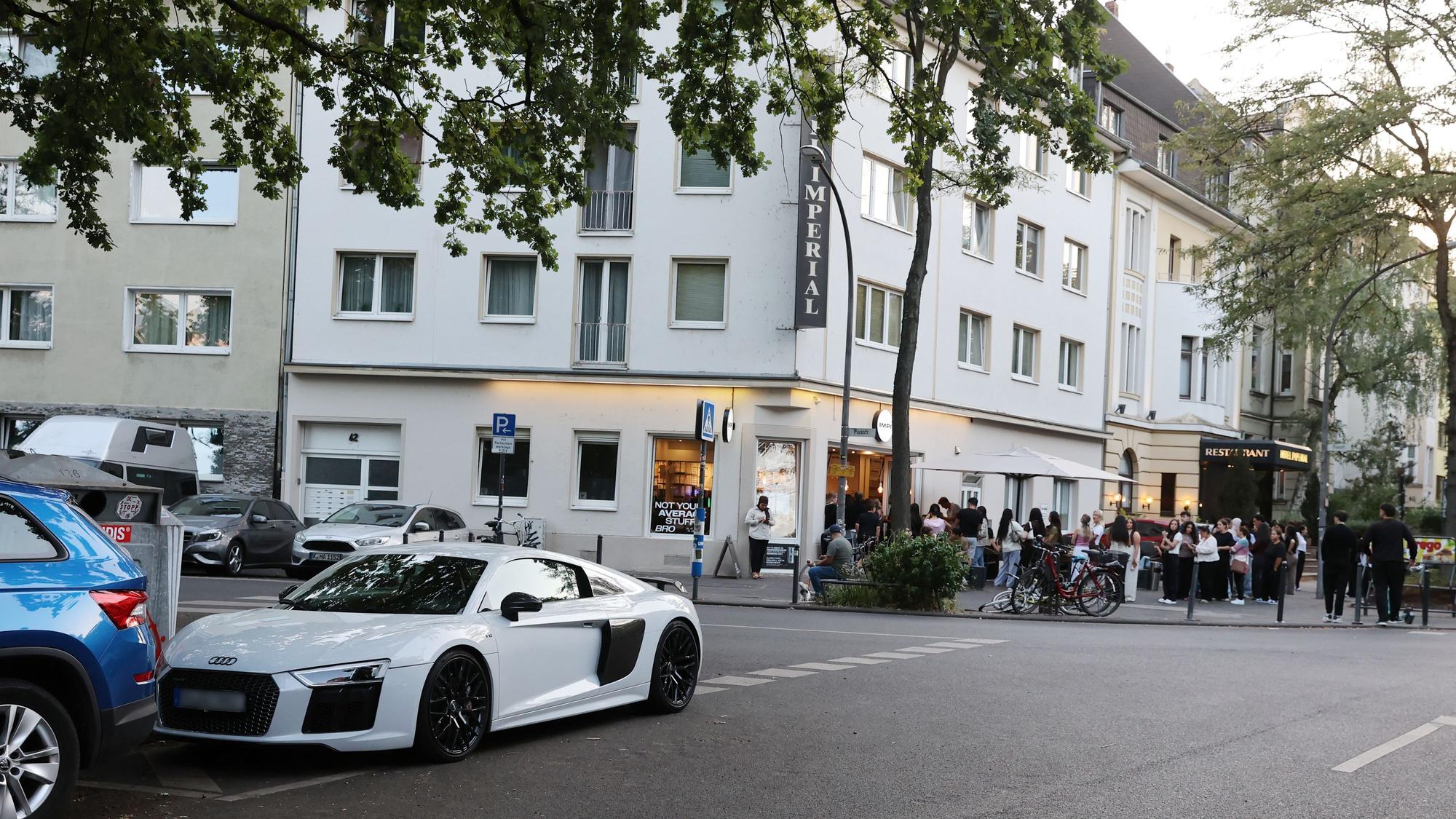 An einer Straßenkreuzung stehen Menschen Schlange vor einem Café. An der Ecke parkt ein PS-starkes Auto verkehrswidrig.