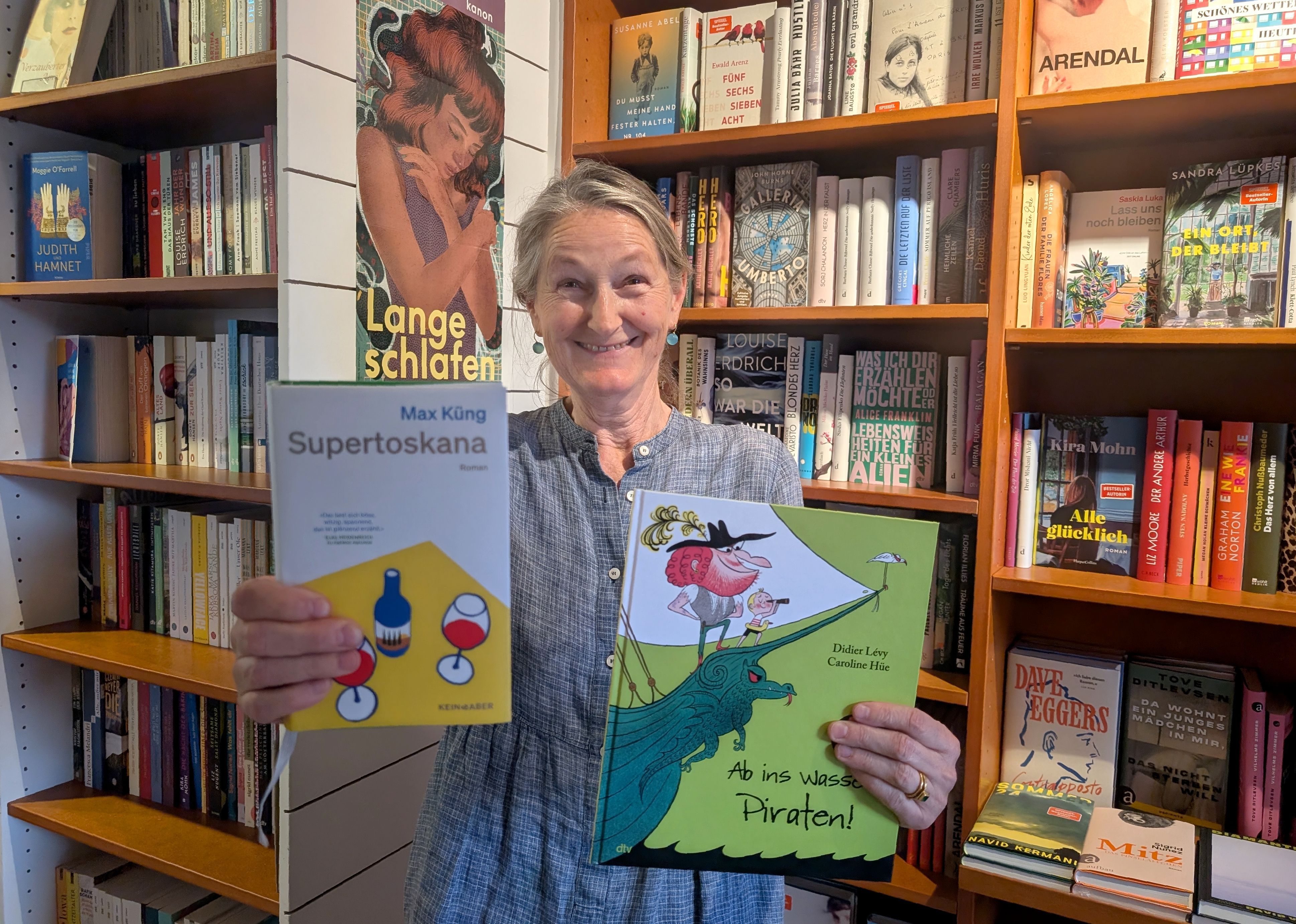Katharina Pütz steht vor einem Bücherregal und hält zwei Bücher in den ausgestreckten Händen.