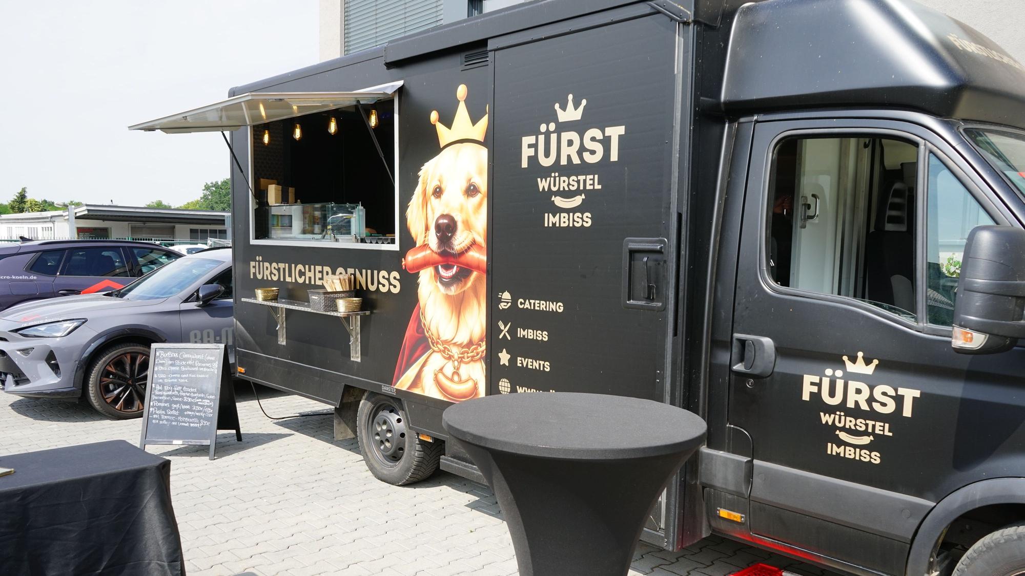 Zu sehen ist ein schwarzer Foodtruck mit einem KI-generierten Bild von einem Hund mit Wurst und Krone darauf.