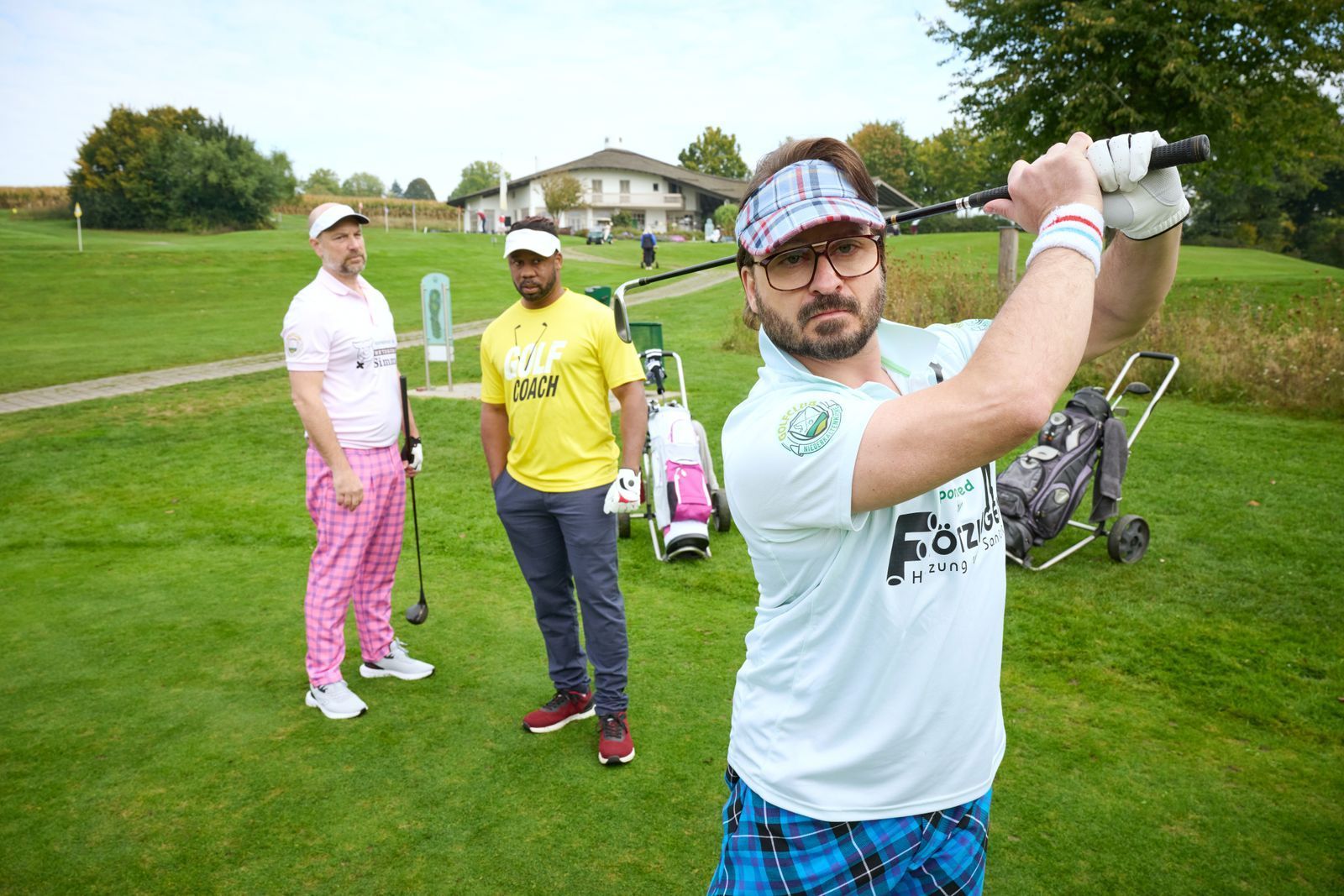 Simmerl (Stephan Zinner, l.) und Flötzinger (Daniel Christensen, r.) versuchen sich unter Aufsicht von Golflehrer Buengo (Castro Dokyi Affum) auf dem Grün.  (Bild: Constantin Film Distribution GmbH / Bernd Schuller)