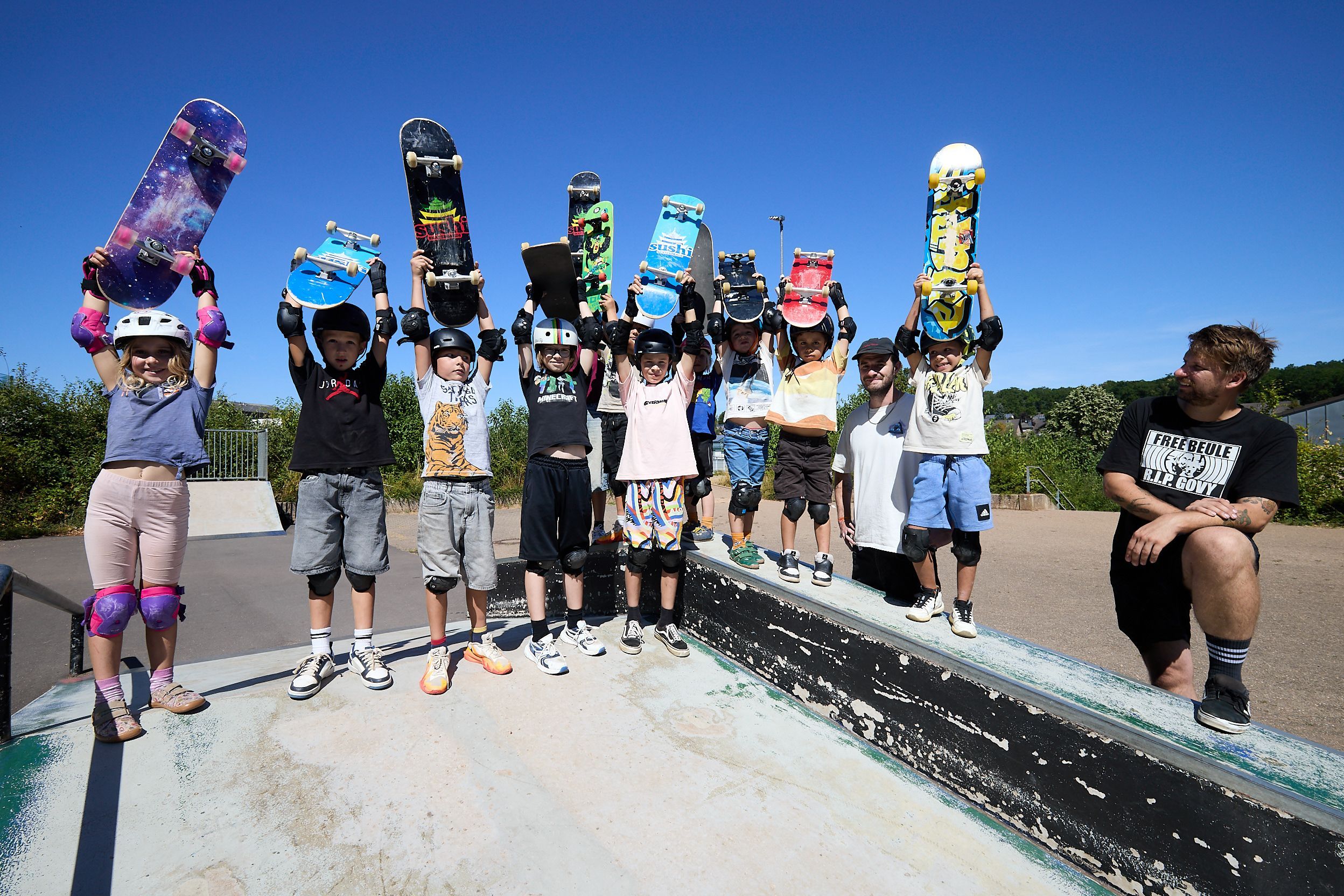 Zehn Kinder halten ihre Skateboards in den Himmel. Zwei Männer schauen zu.