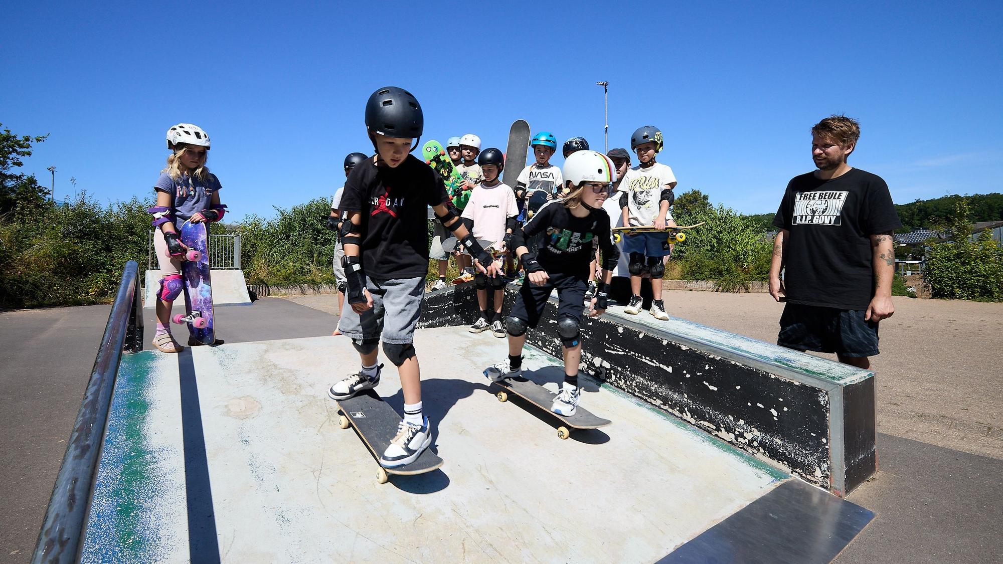 Zwei Kids fahren mit ihren Boards eine Rampe herunter. Die anderen Kinder und ein Mann schauen zu.