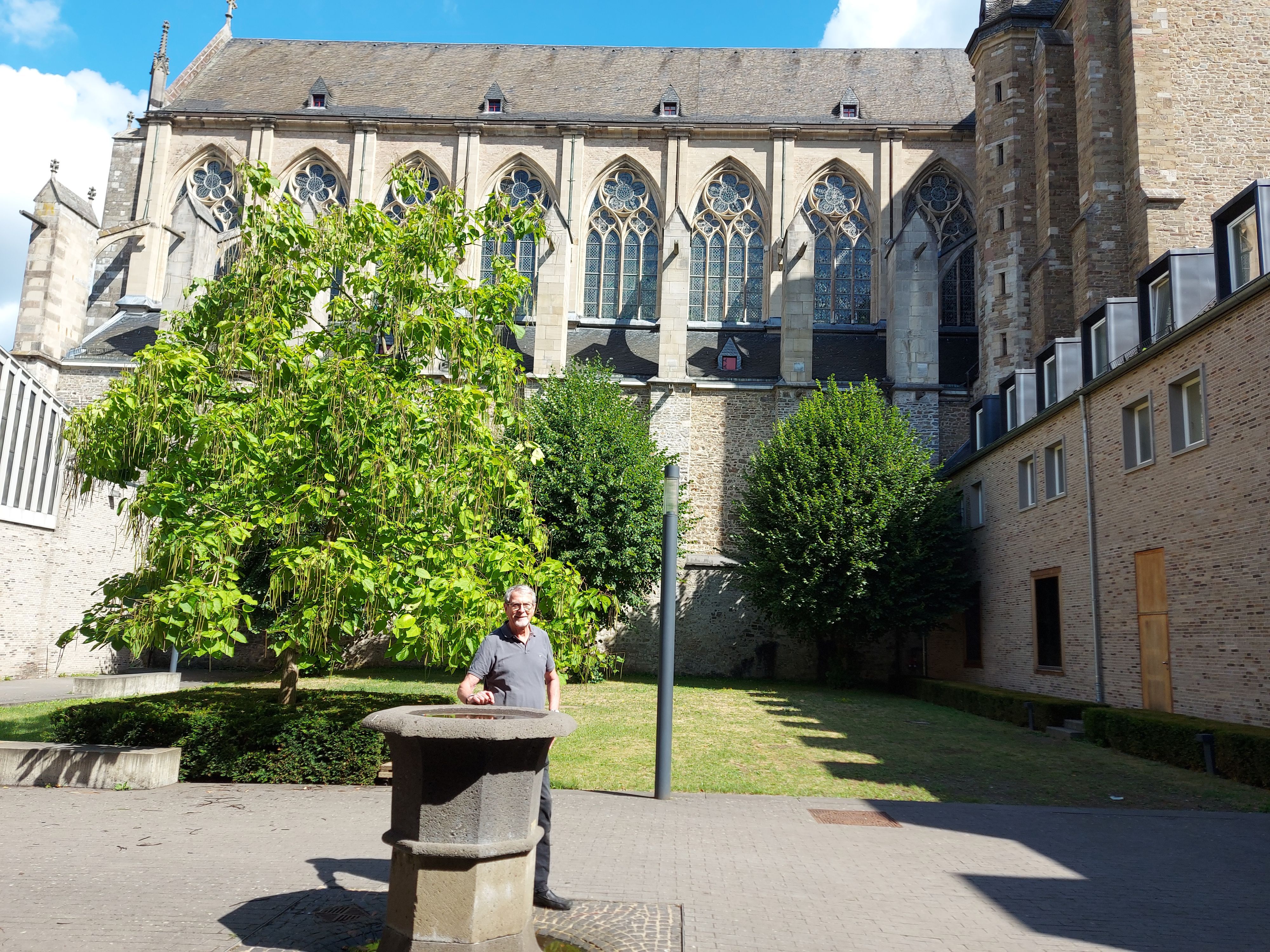 Ein Mann steht an einem steinernen Kübel, im Hintergrund die Fenster einer gotischen Kirche.