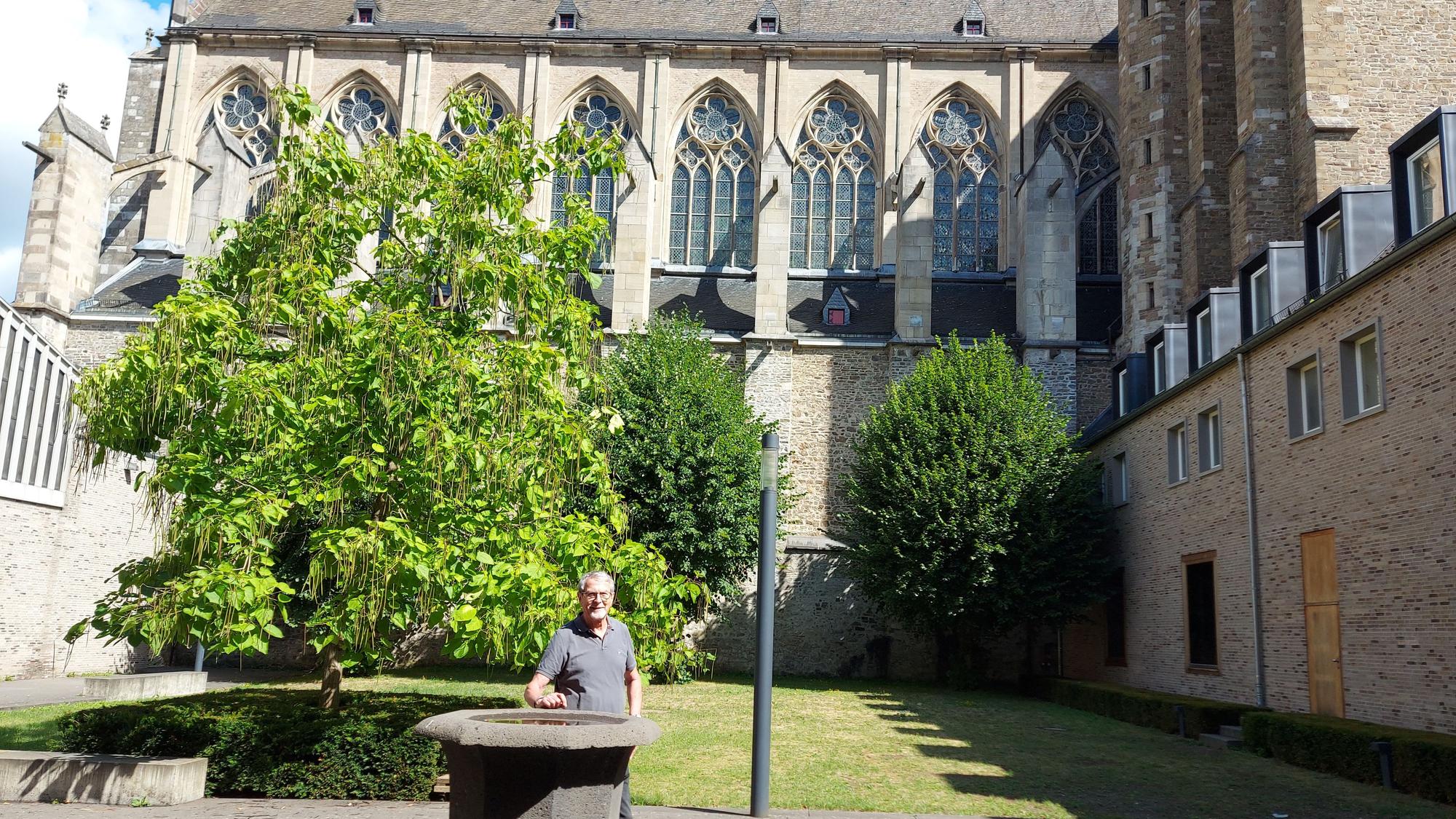 Ein Mann steht an einem steinernen Kübel, im Hintergrund die Fenster einer gotischen Kirche.