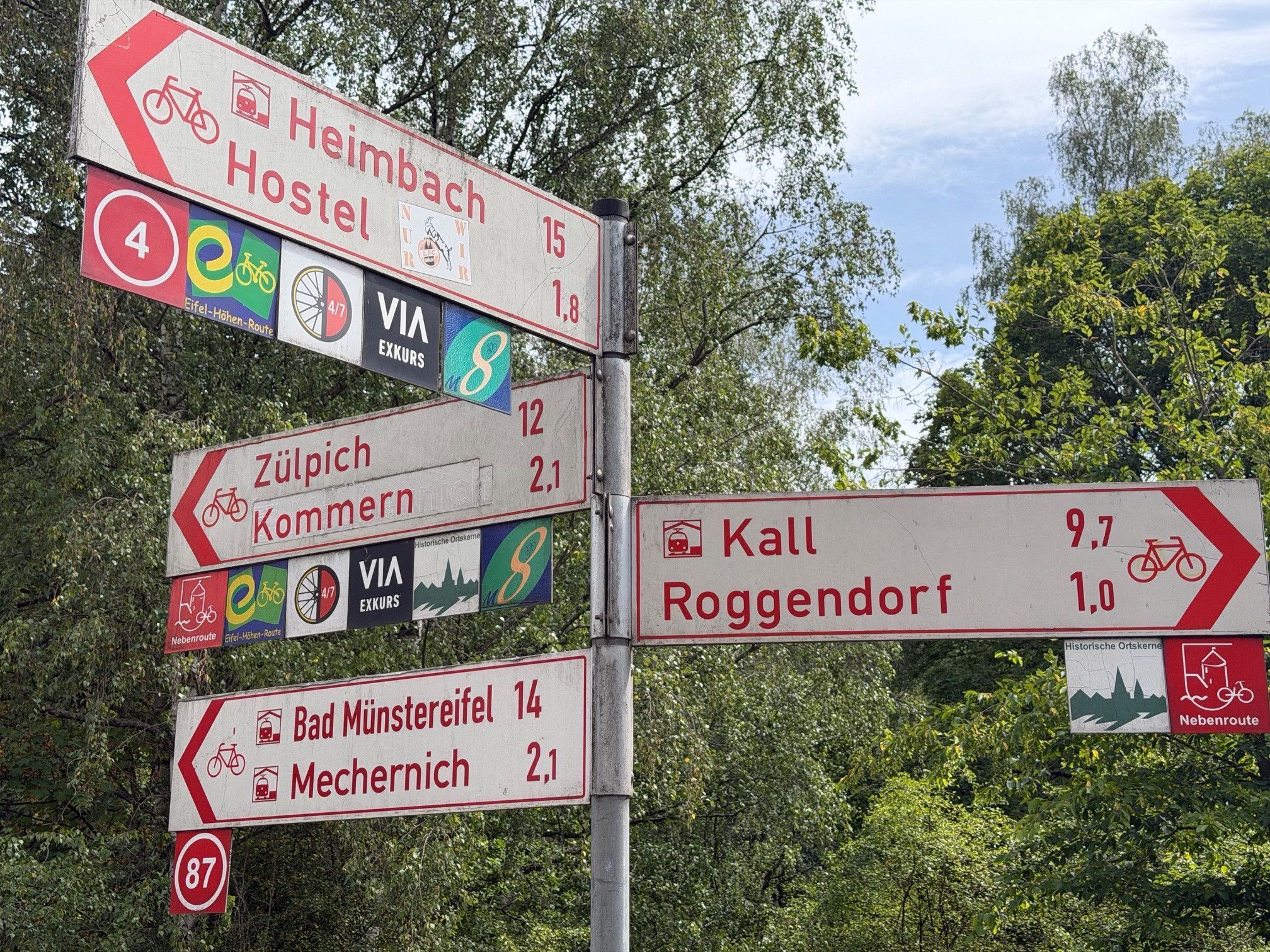 Vier weiße Hinweisschileder mit roter Schrift weisen in unterschiedliche Richtungen.