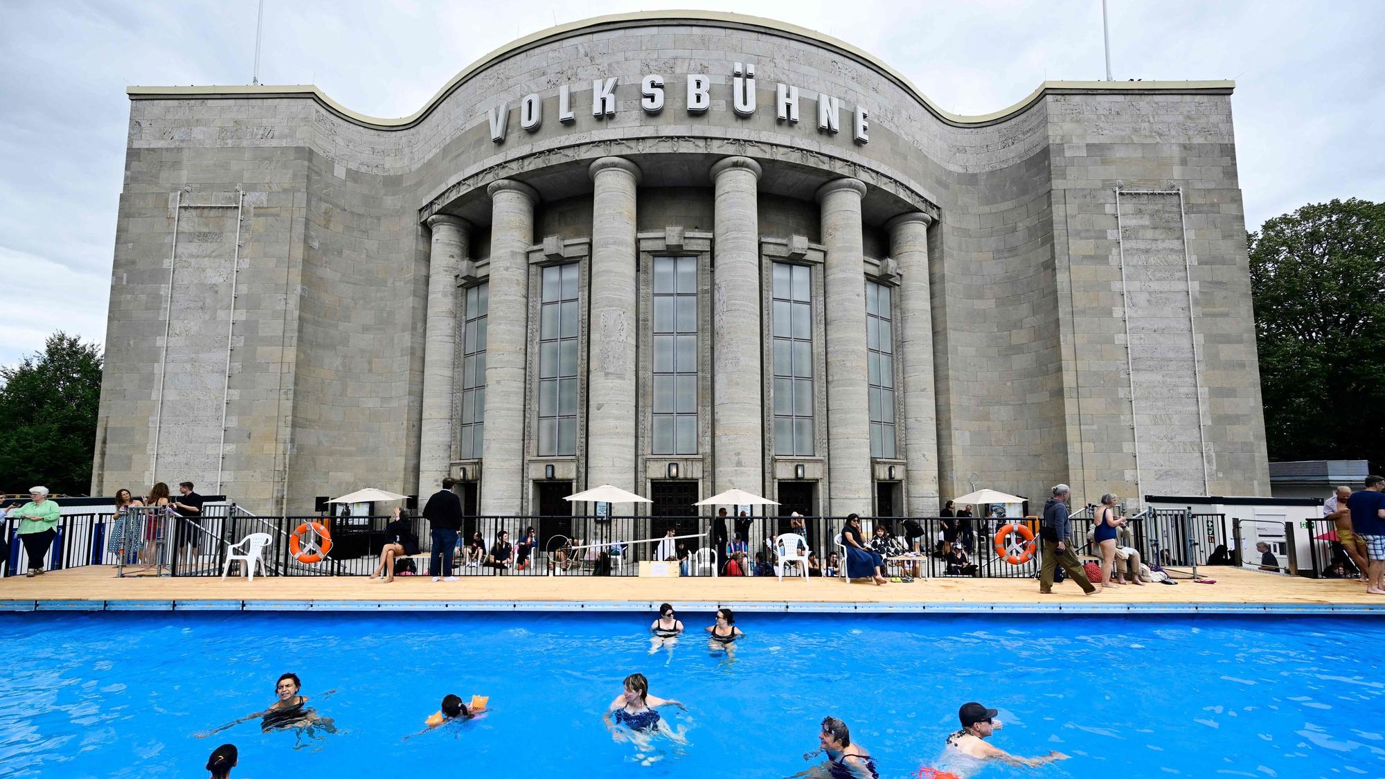 Vor einem Gebäude ist ein großer Pool aufgebaut, in dem Menschen schwimmen.