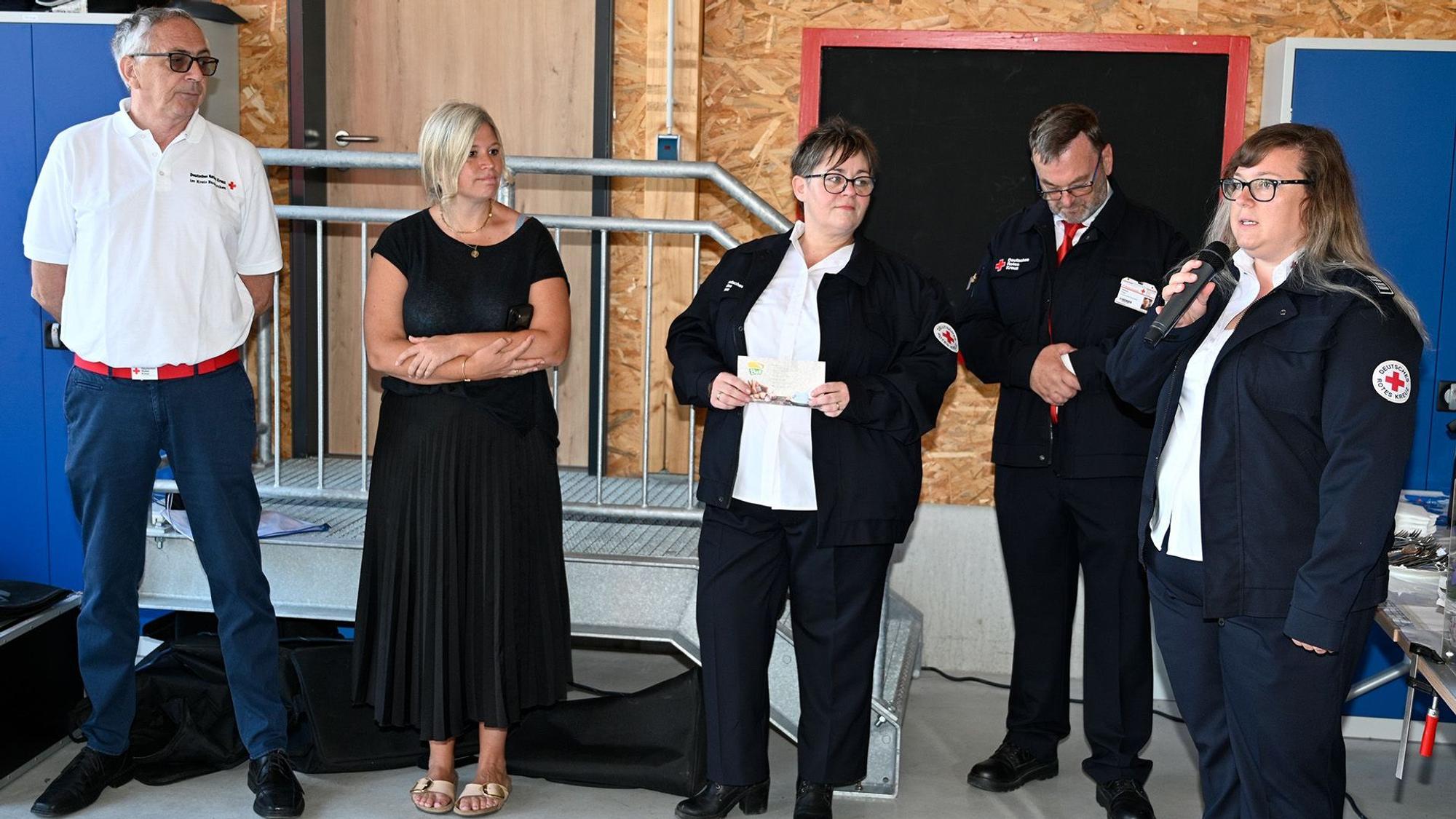 Eine Frau in Rotkreuz-Weste spricht in ein Mikro, zwei Männer und zwei Frauen stehen neben ihr.