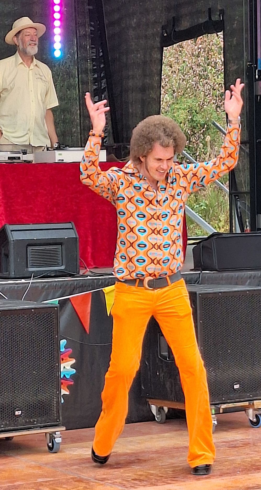 Ein Mann mit Lockenfrisur, buntem Hemd und orangefarbener Hose hat die Arme beim Tanzen erhoben.