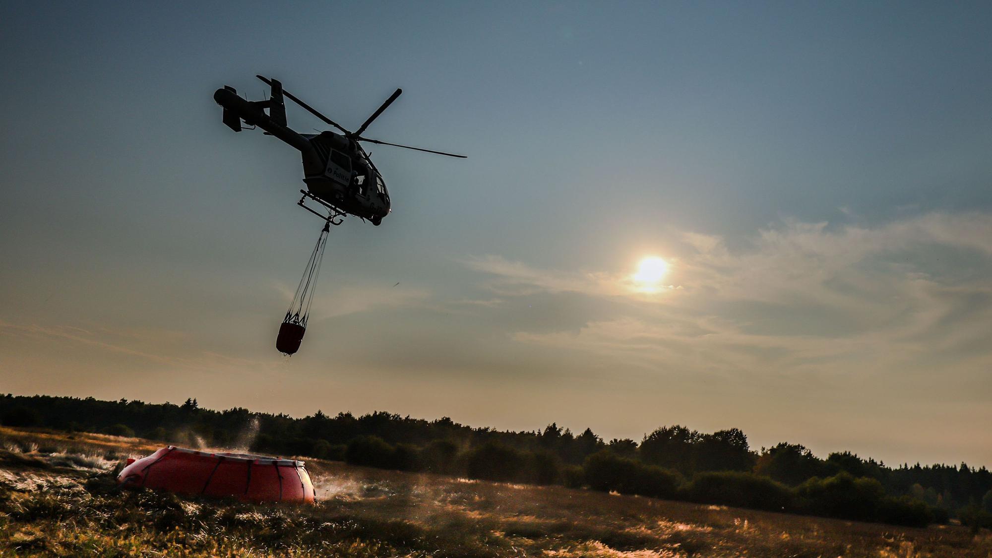 Ein Hubschrauber der belgischen Polizei mit einem Bambi Bucket, ein Löschwasser-Außenlastbehälter, fliegt zu einer provisorischen Wasseraufladestelle im Hohen Venn, um Löschwasser aufzutanken.