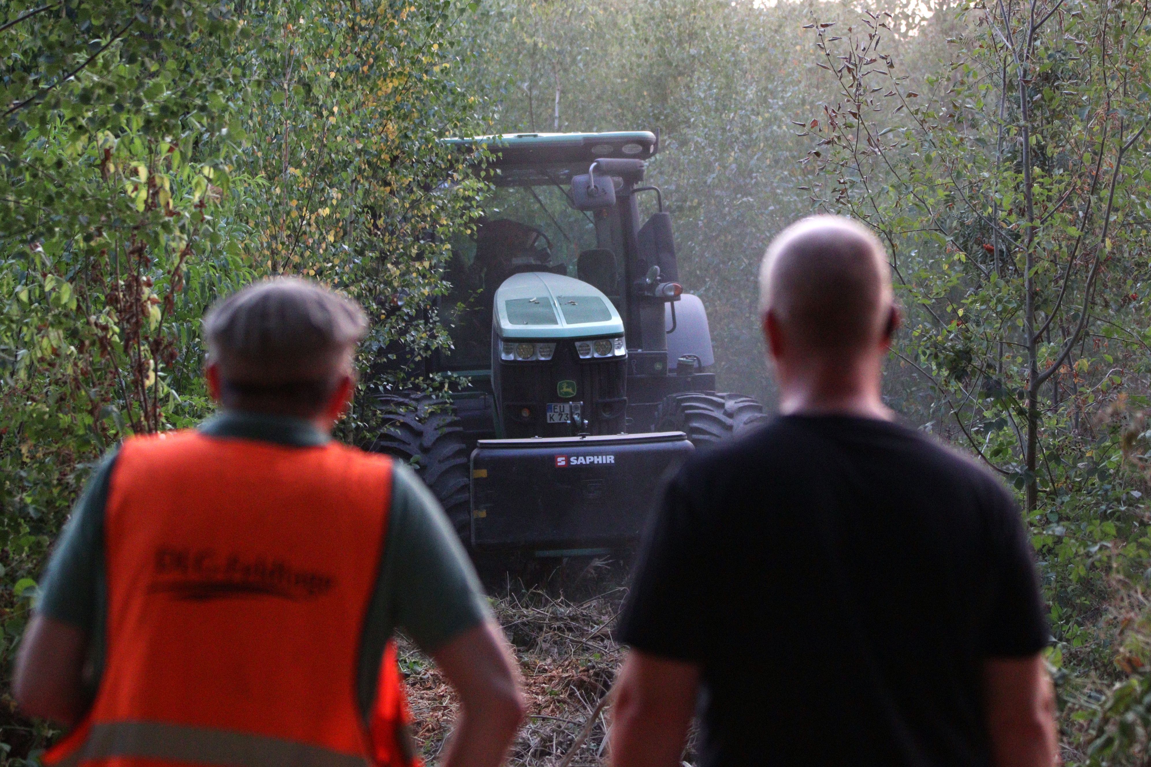 Zwei Personen schauen zu einem Traktor, der in einem Waldstück steht.