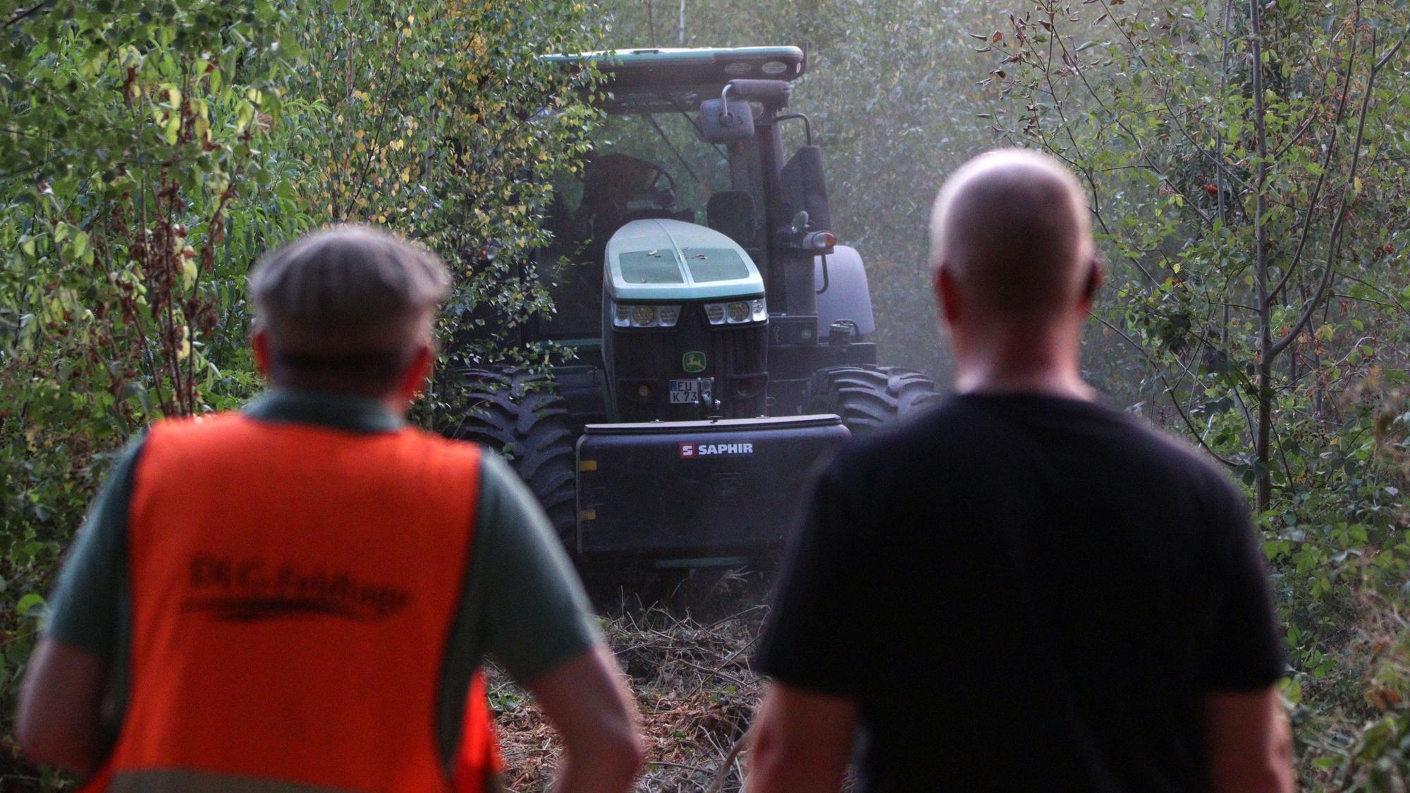 Zwei Personen schauen zu einem Traktor, der in einem Waldstück steht.