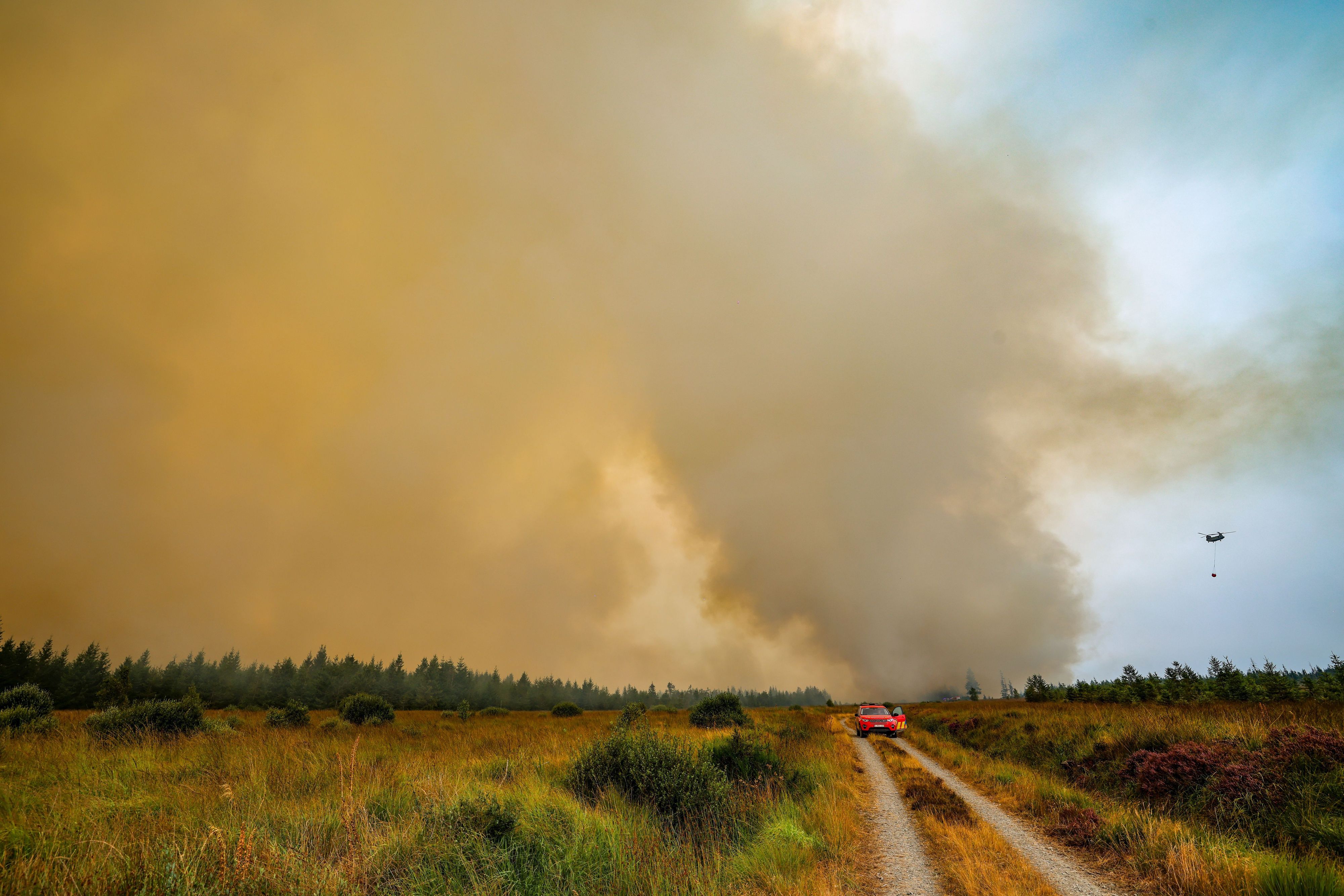 Die belgische Feuerwehr bekämpft den Brand in der Hochmoor- und Sumpflandschaft Hohe Venn unweit der deutschen Grenze. Gleichzeitig ist ein Chinook-Hubschrauber mit einem Wassersack zur Bekämpfung der Flammen im Einsatz.