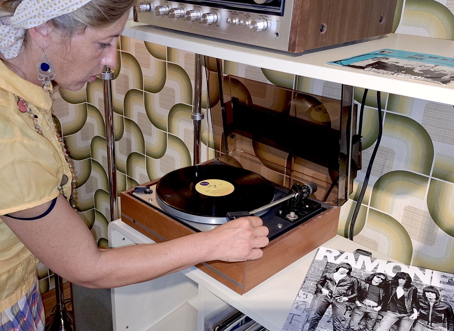 Eine Frau spielt eine LP der Ramones auf einem Dual-Plattenspieler ab, der in einem Regal vor einer buntgemusterten Tapete steht.