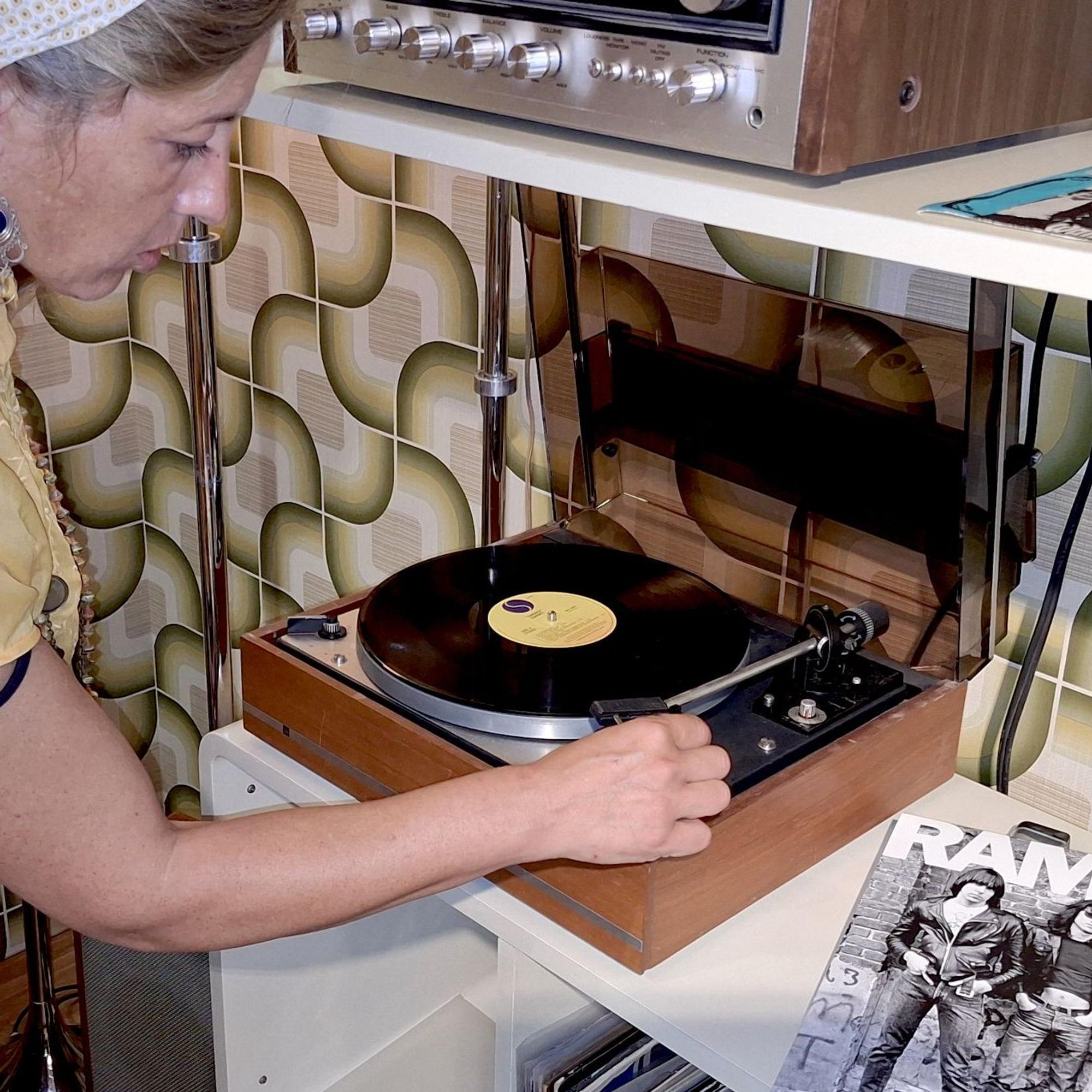 Eine Frau spielt eine LP der Ramones auf einem Dual-Plattenspieler ab, der in einem Regal vor einer buntgemusterten Tapete steht.