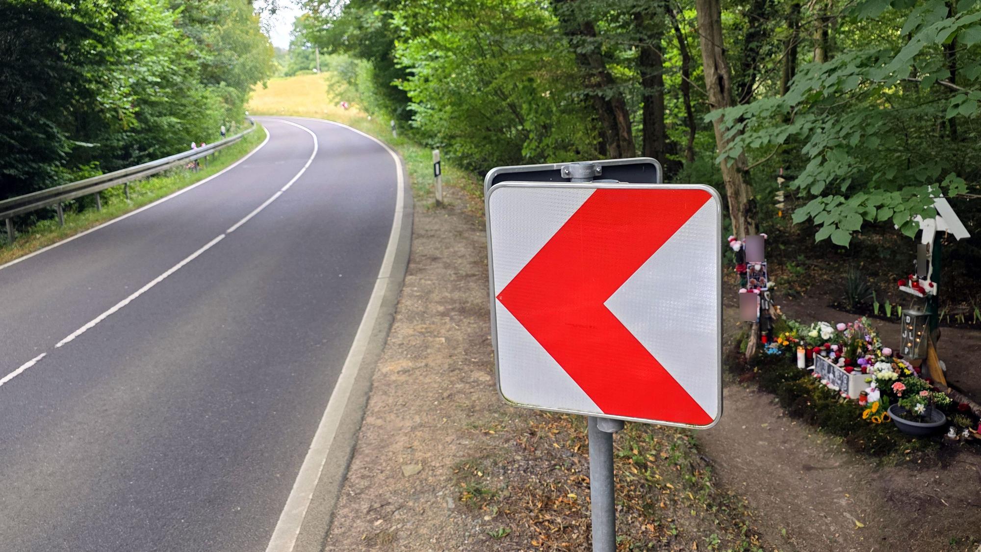 Eine Warntafel, eine Kreuz und Blumen stehen neben der Landstraße im Dürschtal bei Kürten-Dürscheid, an der eine Leitplanke fehlt.