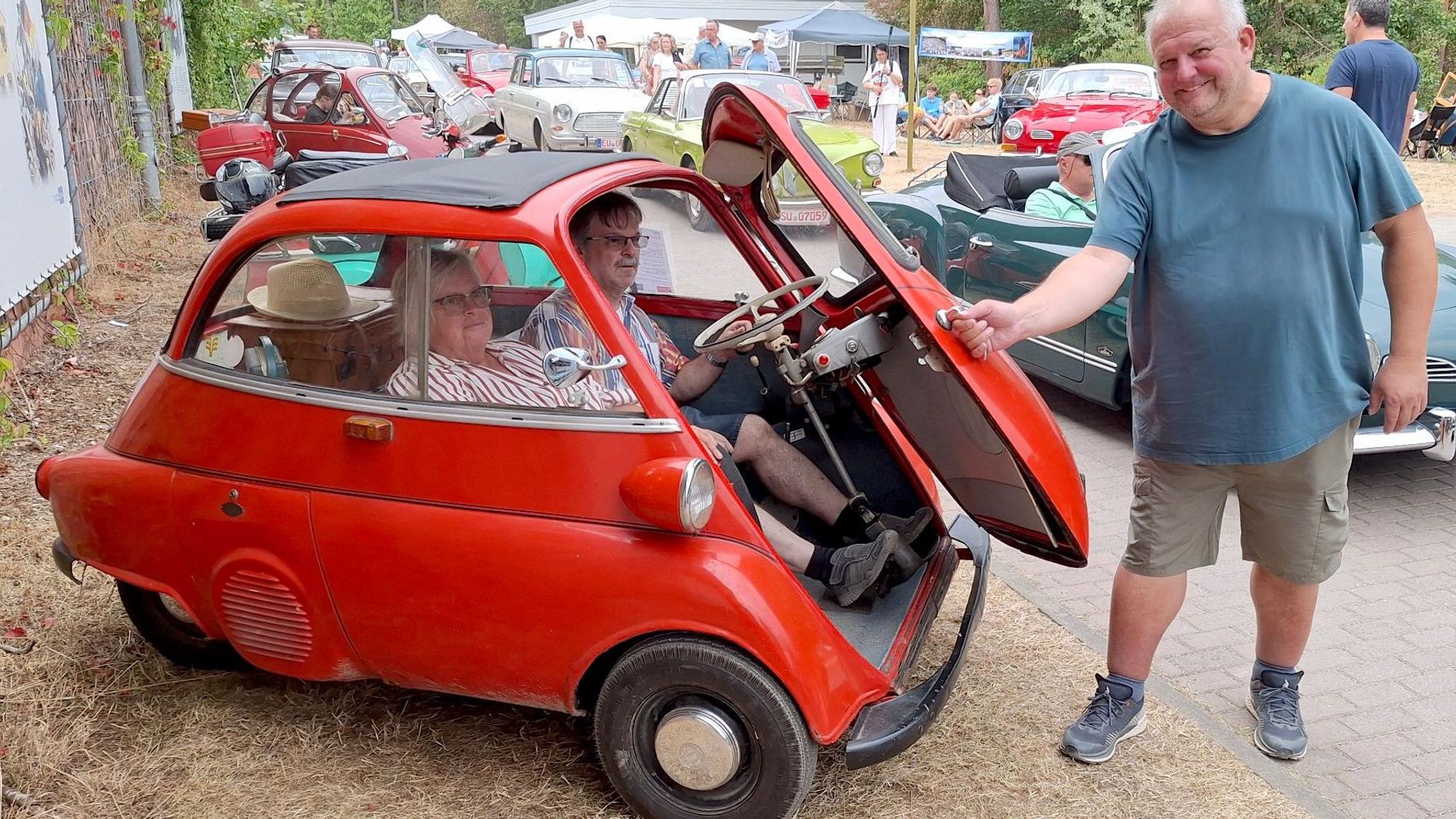 Ein Mann schließt die Klappe einer roten BMW Isetta, in der zwei Personen sitzen.