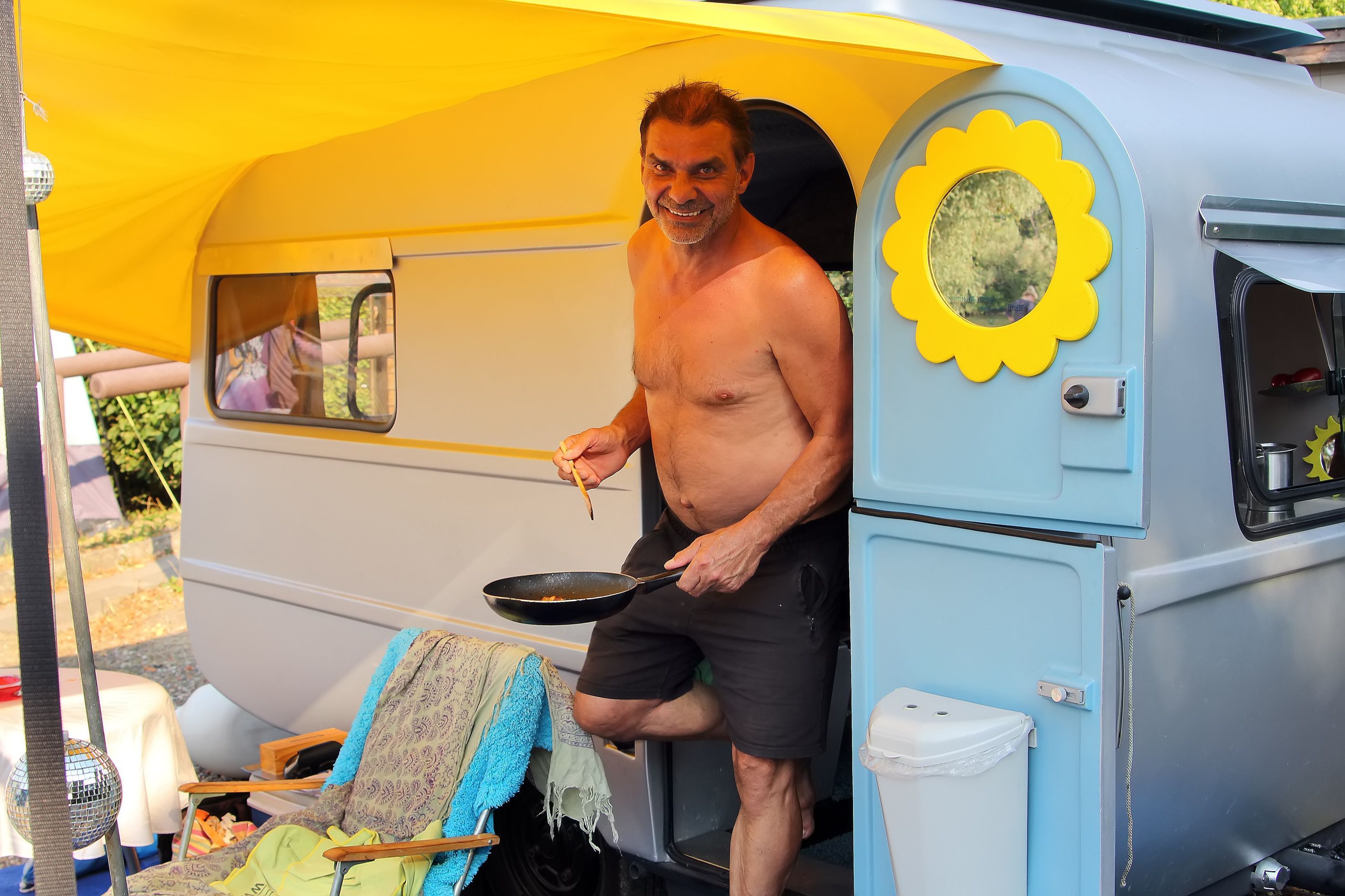 Das Bild zeigt einen Mann mit Pfanne in der Hand an seinem Campingwagen.